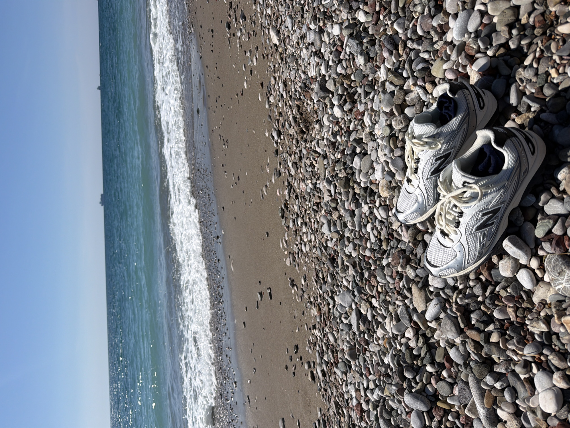 A pair of white and silver New Balance sneakers sits on a pebble beach. The sea is to the left, with waves lapping the shore. The sky is light blue. The beach is in Bar, Montenegro.