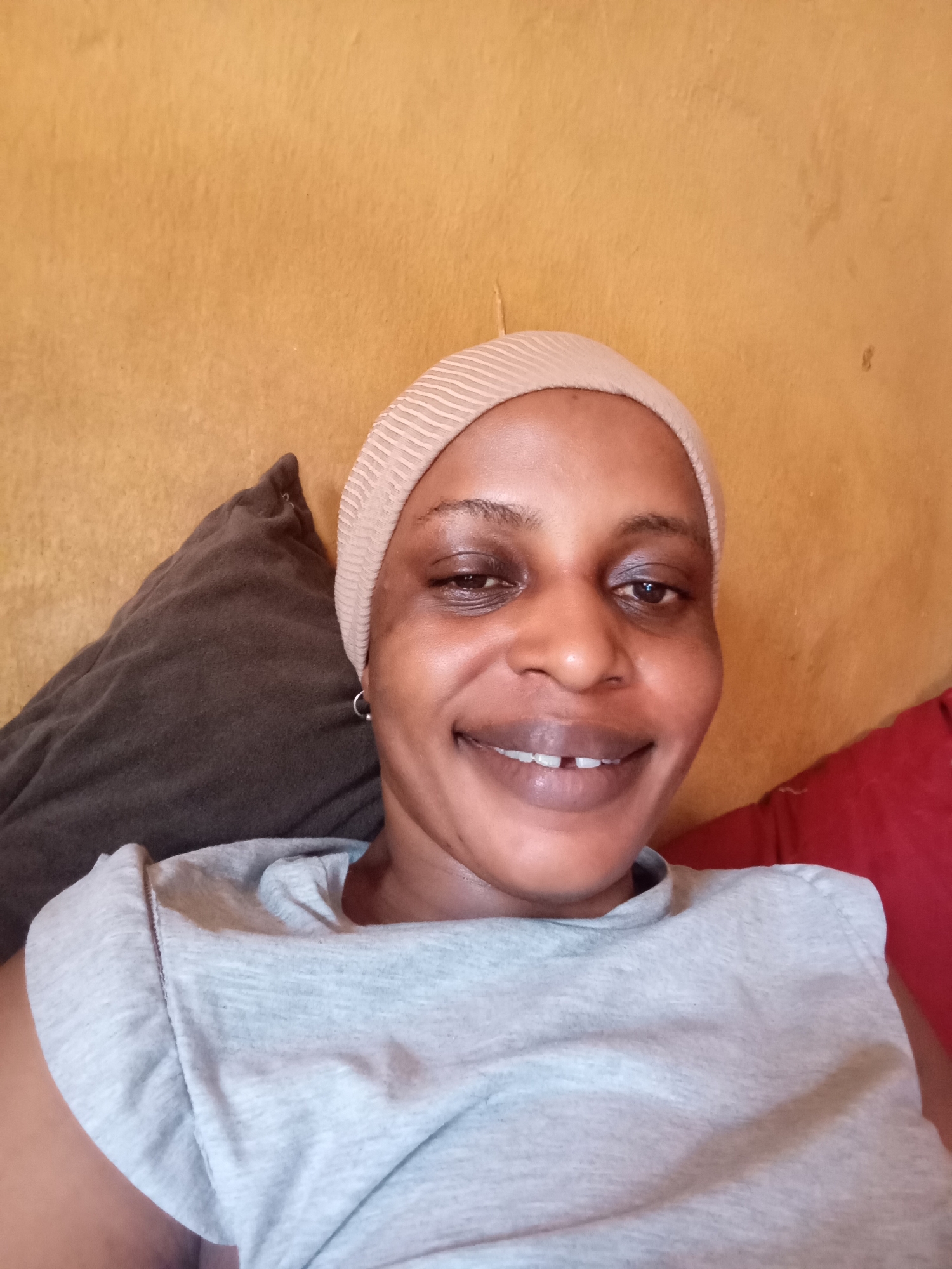 A head-and-shoulders selfie of a smiling Black woman is centered in the frame. She wears a beige, striped head covering and a light grey t-shirt. The woman has dark eyes, and a gap in her upper front teeth is visible. She is angled slightly to the left, with her face towards the camera. Her left ear is visible, displaying a small silver hoop earring. Behind her is a textured yellow wall, with a dark grey pillow on the left side of the frame and a red cushion on the right. Based on the information provided, this photo was taken in Narayi, Nigeria.