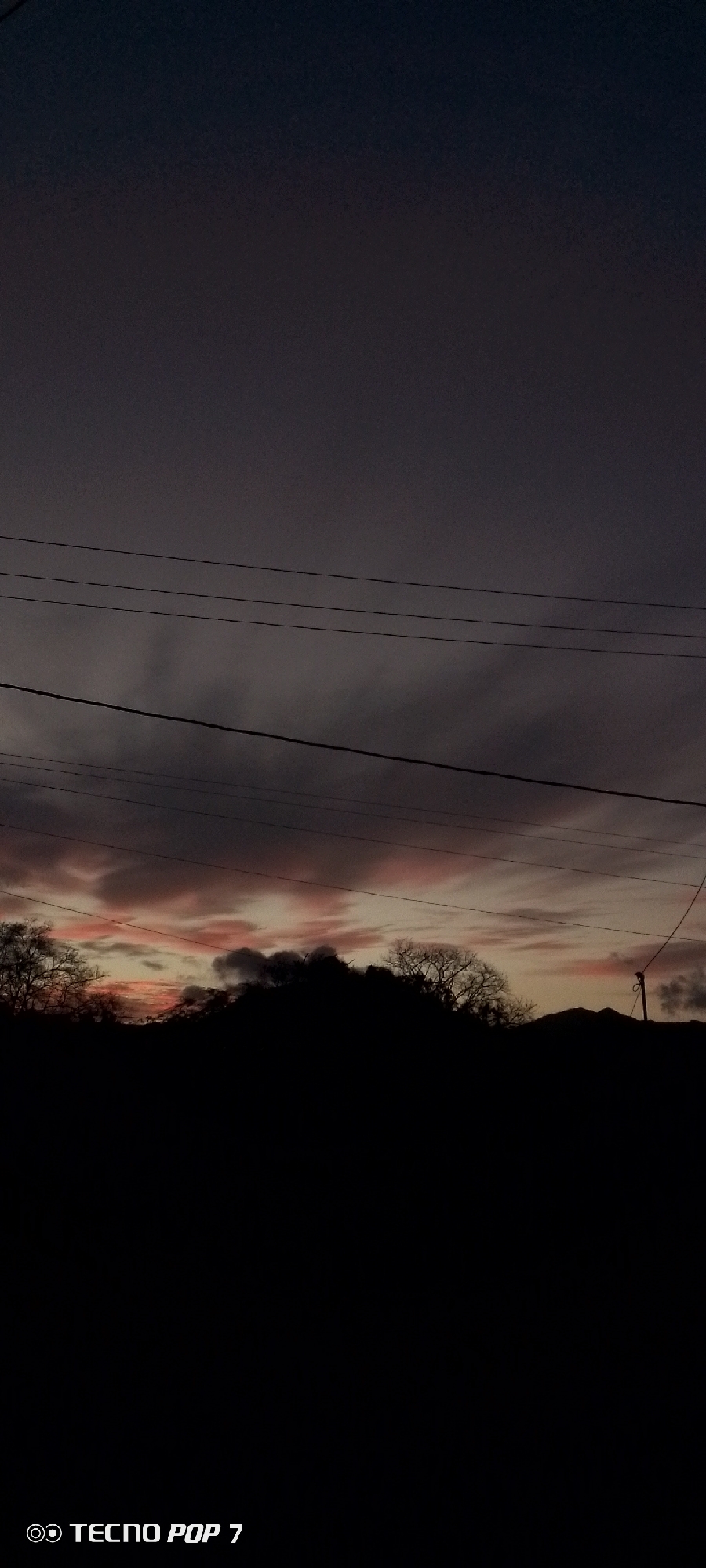 The image depicts a dusk or dawn scene in Altagracia, Venezuela. The sky is predominantly dark with subtle pink and orange hues peeking through the clouds near the horizon. Silhouetted trees and hills form a dark, irregular line across the lower third of the frame. Utility lines run horizontally across the middle portion of the sky. The lower portion of the image is entirely dark. "TECNO POP 7" is imprinted at the bottom left.
