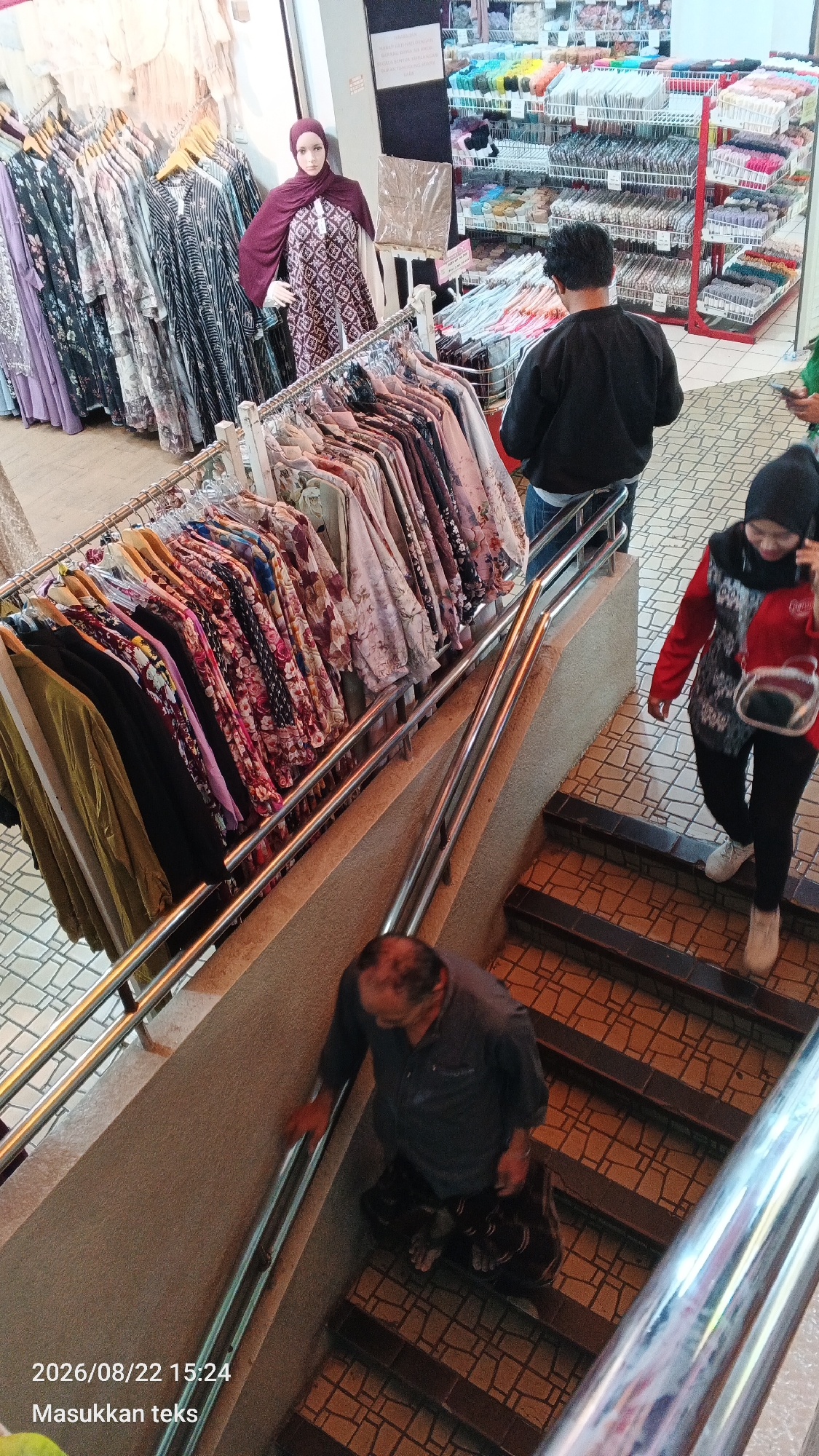 This image captures a bustling scene inside a clothing store, possibly located in Malang, Indonesia, as indicated by the timestamp and text overlay. The store features a wide array of garments displayed on racks, with a particular emphasis on women's clothing featuring floral patterns and modest designs, suggesting a focus on Islamic fashion.
In the foreground, a man is descending a staircase, his hand resting on the metal railing. He is wearing a dark blue long-sleeved shirt and patterned trousers. A woman in a red and black top, black pants, and a black hijab is also visible on the stairs, walking upwards and carrying a red bag. Another person, a man wearing a black jacket and jeans, stands near a clothing rack, appearing to browse. A mannequin dressed in a burgundy hijab and a patterned dress is prominently displayed near the clothing racks, serving as a visual representation of the store's offerings.
The store's interior is characterized by tiled flooring and metal railings, creating a functional and perhaps multi-level shopping environment. Shelves filled with various items, possibly fabric or accessories, are visible in the background. The overall impression is one of a busy retail space catering to a diverse clientele. The timestamp and text "2026/08/22 15:24 Masukkan teks" suggest this image was taken on August 22, 2026, at 3:24 PM, and may have been intended for further annotation or labeling.