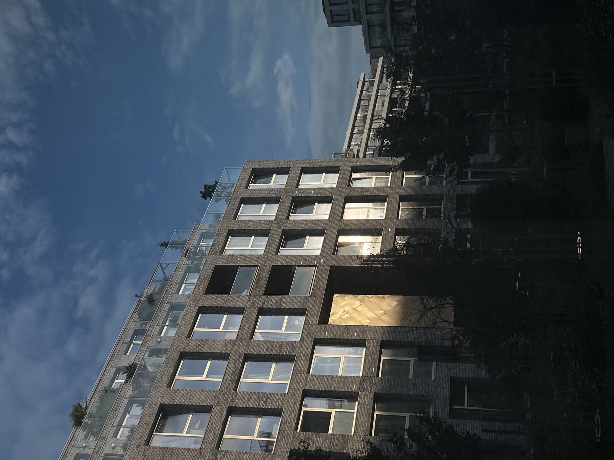 The image shows a modern apartment building with a gray brick facade and large windows. The building has multiple balconies with glass railings, and some greenery is visible on the balconies. The sky above is blue with some scattered clouds. In the foreground, there are dark silhouettes of trees, suggesting a park or garden area in front of the building. The lighting suggests it is either early morning or late afternoon, with sunlight hitting one side of the building and casting shadows. The architectural style and the presence of water in the background (not clearly visible but implied by the context of Amsterdam) suggest this could be a residential area in Amsterdam.