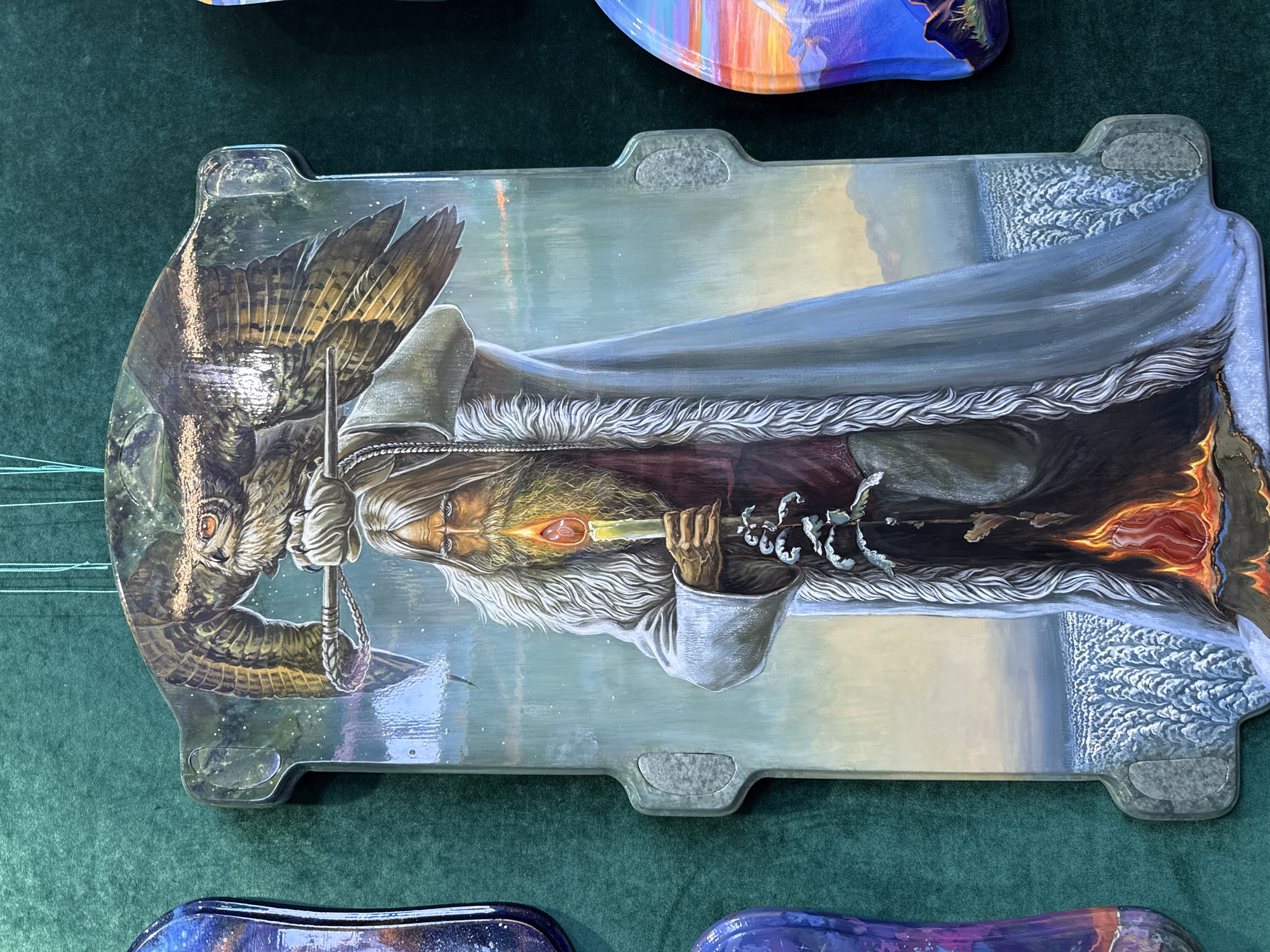 A rectangular, possibly hand-painted, decorative object depicting a wizard, similar in appearance to Gandalf from the Lord of the Rings, with an owl, on a dark green felt surface. The wizard has a long white beard and hair and wears a long grey cloak over a burgundy garment. He holds a staff with a flame-like object at its tip. The owl is perched beside him. The object has decorative tabs on its sides. The background is a muted, possibly painted scene. The location, Upper Uymon, Russia, is not evident from the image.