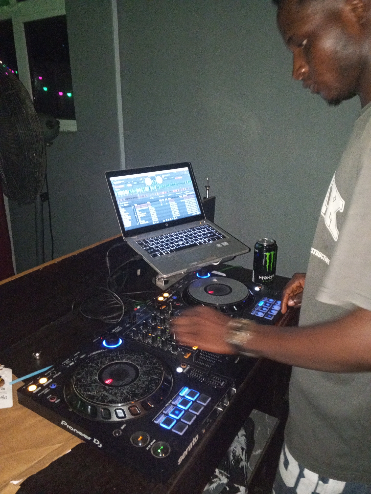The image shows a DJ, likely male, operating a Pioneer DJ Serato mixing console connected to a laptop displaying DJ software. A can of Monster energy drink sits next to the console. The scene is indoors, dimly lit, potentially a club or event space. A fan is visible in the background, and there are multi-colored lights through a window, providing additional visual context for an entertainment setting. Without additional context, the location in Lokoja, Nigeria cannot be verified, but the setting implies an urban entertainment environment.