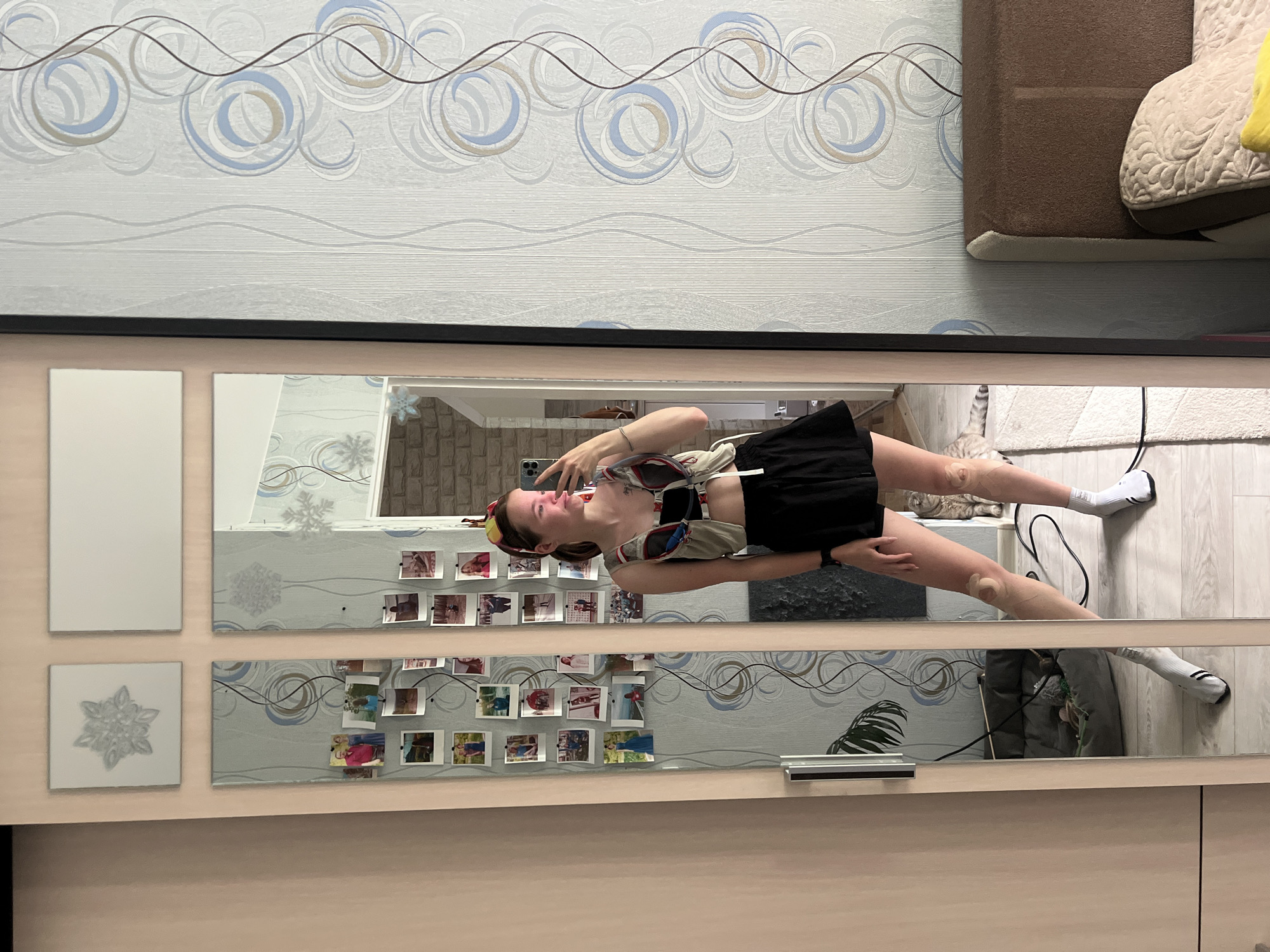 A young woman is taking a mirror selfie in a bedroom. She is wearing a black skirt, white socks, and a grey and red vest. The room has patterned wallpaper and a large mirror on the wall. A cat is visible in the reflection of the mirror. There are also several framed photos on the mirror. The woman is standing in front of a bed.