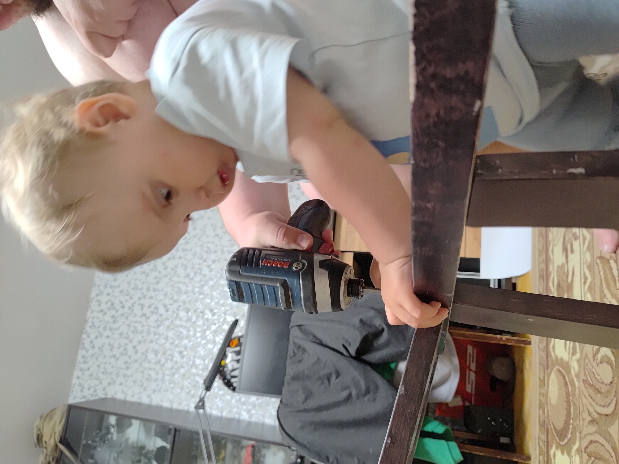 A toddler, with blonde hair and wearing a white shirt, is holding a Bosch cordless impact driver (model GDR 10.8-LI) and seemingly attaching a wooden chair leg with it. An adult is positioned behind the toddler, possibly assisting or supervising. The scene is indoors, with a patterned wallpaper visible in the background. A glass cabinet and a floor lamp are visible. Part of a patterned rug or carpet is also in view. The location information (Марковский, Russia) is provided but not discernible from the image itself.