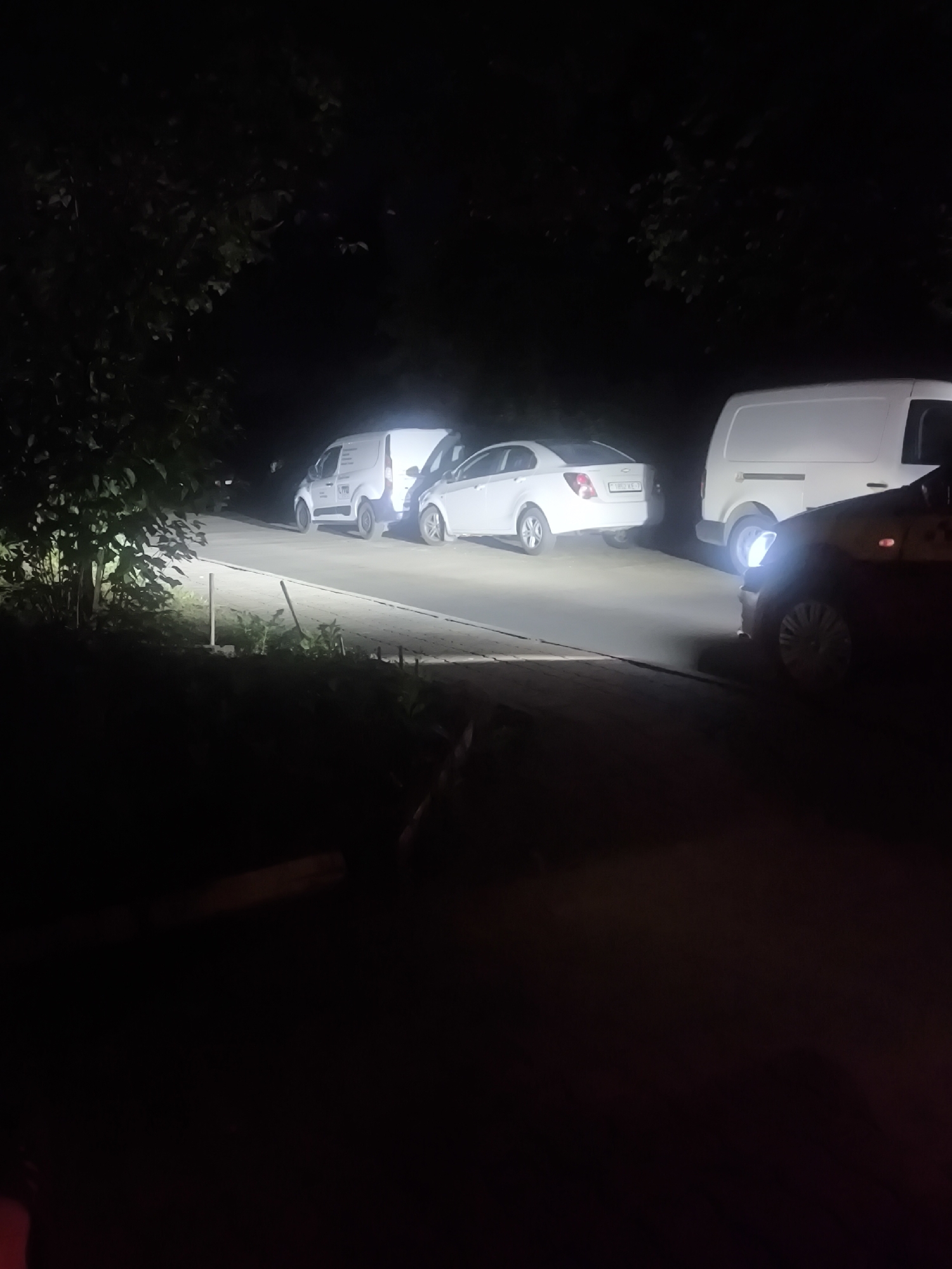 At night, on a paved road in Minsk, Belarus, a white Chevrolet Aveo is parked, its rear facing the camera. To its right is a white utility van, also parked. To the right of the van, a dark-colored car is parked, its headlights illuminating the scene. The area is bordered by trees and foliage.