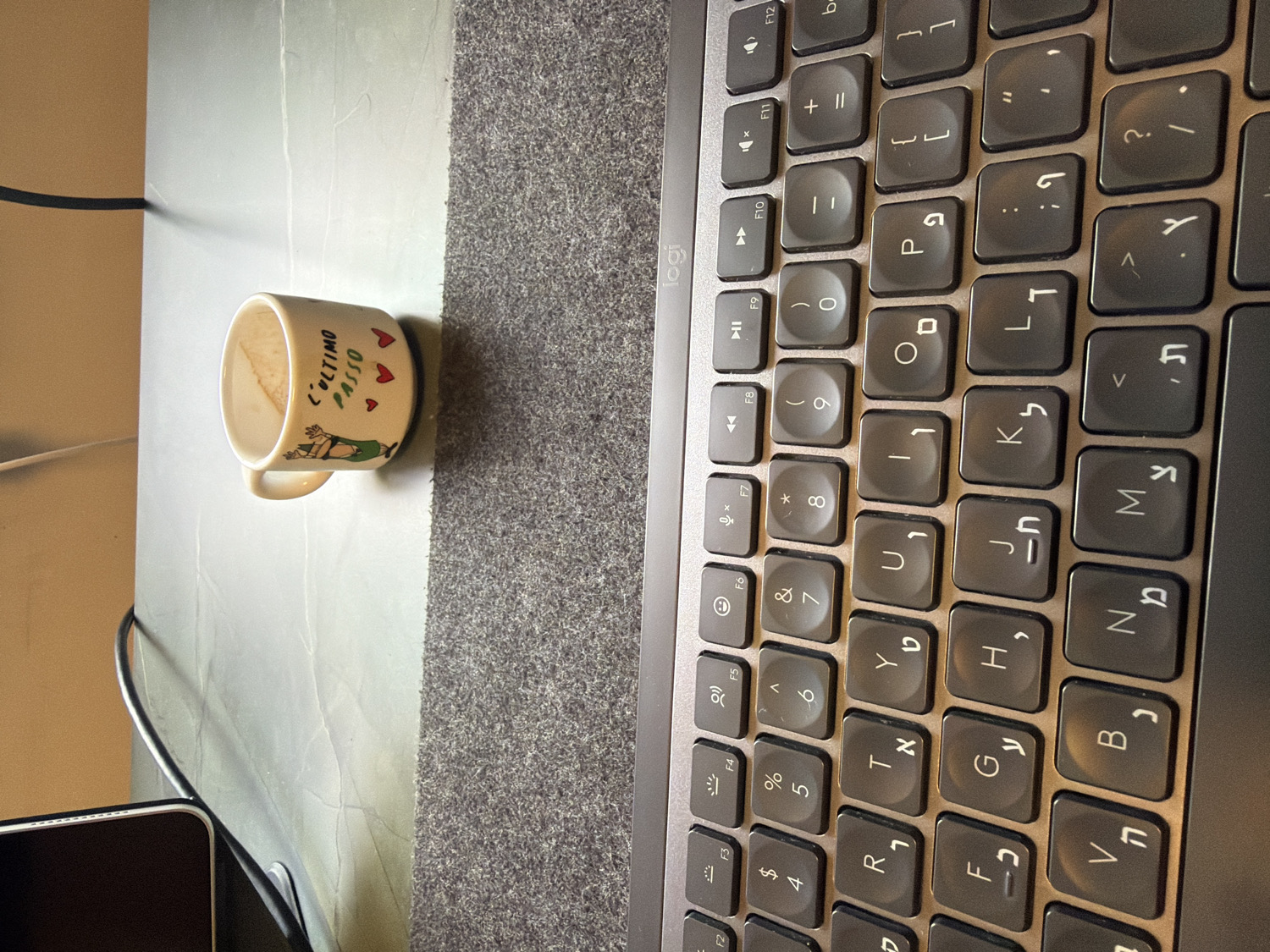 An overhead view captures a workspace, primarily featuring a laptop keyboard and a ceramic mug, resting on a surface partly covered by a grey textured mat.
The main subjects are a dark grey laptop keyboard from the brand "Logi" (Logitech) and an off-white cylindrical ceramic mug with a handle. The keyboard features a bilingual layout with both Latin and Hebrew characters, indicating its use in a region like Kafr Yasif, Israel. Several function keys (F1-F12) are visible, along with alphanumeric keys, punctuation, and symbols. The mug has brown residue inside, suggesting it recently held a beverage like coffee or tea.
On the side of the mug, there is a cartoon drawing of a reclining figure and the Italian text "L'ULTIMO PASSO" (meaning "the last step" or "the final stride"), accompanied by three red heart symbols.
The objects are placed on a dual-textured surface; the laptop sits on the grey textured mat, while the mug is on an adjacent lighter, smoother surface, possibly a desk. Part of a black electronic device (likely a smartphone or another portable device) and a black cable are visible in the bottom left corner, indicating a busy work setup. There are no people visible. The lighting appears to be ambient indoor light.