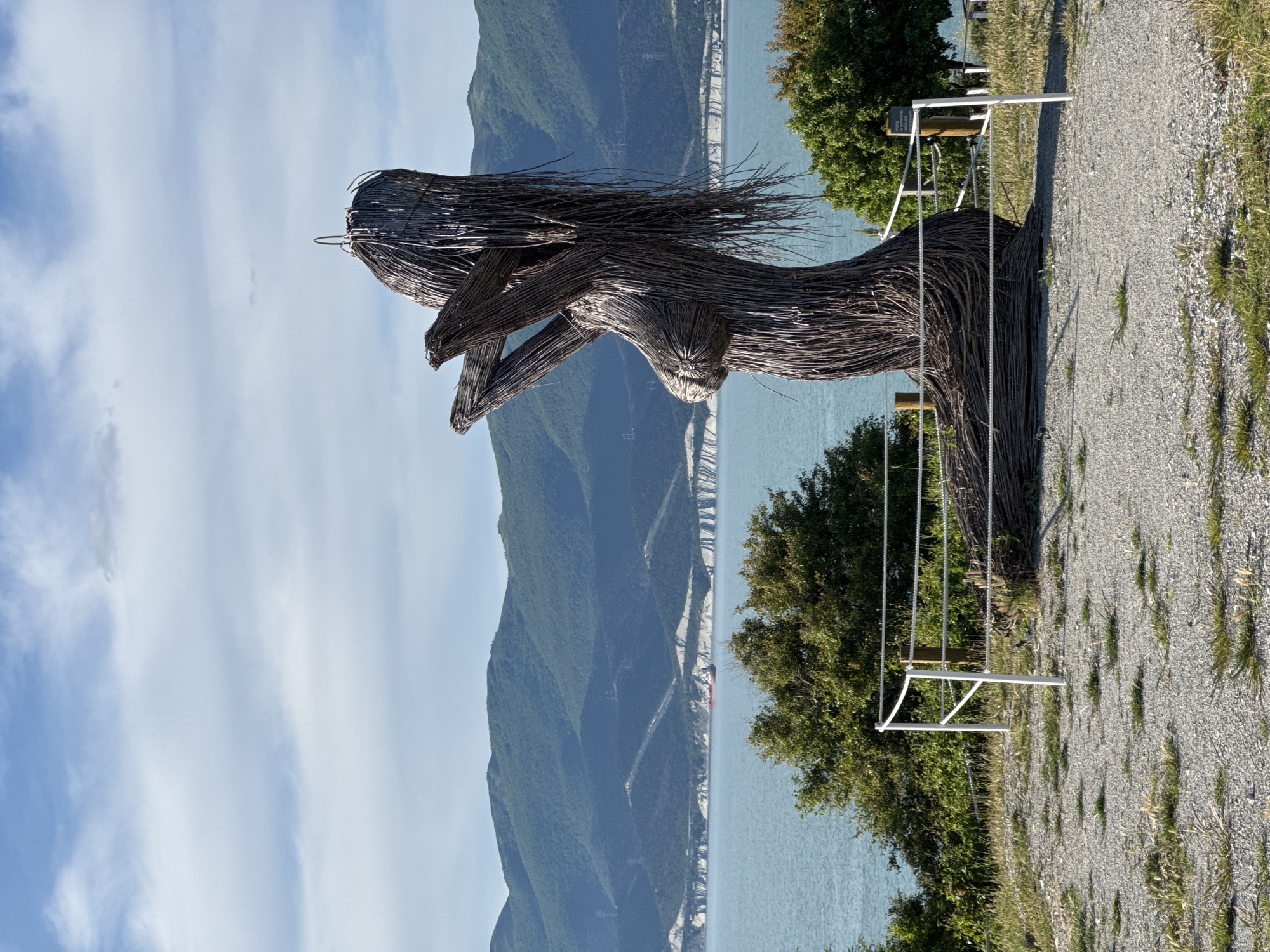 This image presents a large, striking sculpture set against a scenic coastal landscape in Kabardinka, Russia. The central subject is a tall, slender human-like figure, resembling a woman, meticulously crafted from dark, interwoven branches and twigs. The sculpture features long, flowing "hair" made of the same organic material and stands with its arms bent at the elbows, hands held up near the chest in a contemplative or reaching pose.
In the background, a wide expanse of calm, blue-green sea stretches towards a distant horizon. Beyond the water, a series of towering, verdant mountains, densely covered in trees, rise majestically. Along the base of these mountains, white cliffs or rocky formations are visible, creating a stark contrast with the dark green slopes. A small red ship can be spotted in the distant water near the coastline.
The foreground shows a gravel path or ground, suggesting a public viewing area. To the right of the sculpture, green trees and bushes frame the scene. A white metal frame structure, with ropes hanging from it, is visible near the sculpture, indicating a swing set, possibly offering a unique vantage point for the scenic views. A small, dark rectangular sign is attached to this frame, but any text on it is illegible due to distance and resolution.
The time of day is clearly daytime, under a bright blue sky with scattered white clouds, suggesting clear and pleasant weather. The overall scene conveys a sense of natural beauty and artistic integration, with the sculpture serving as a captivating focal point amidst the dramatic coastal scenery. No specific activity is depicted, emphasizing the serene and static nature of the setting.