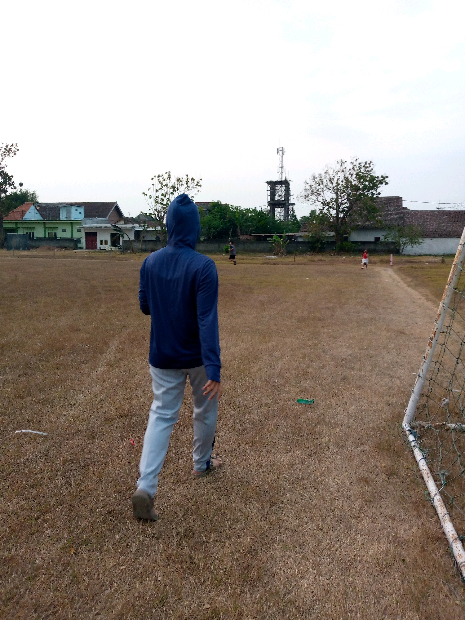 The image depicts a dry, brown grass field with a soccer goal on the right side. A person wearing a dark blue hooded jacket and light gray pants is walking away from the camera towards the center of the field. In the background, there are several houses with red and green roofs, a cell tower, and trees. The sky is overcast. This scene appears to be in a residential area of Pacing, Indonesia, possibly a local sports field.