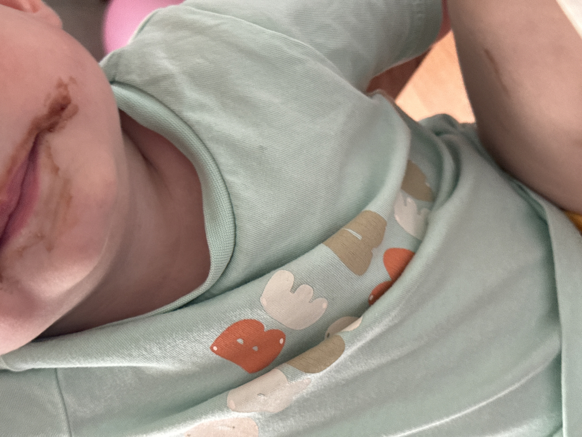 A close-up, eye-level shot captures the chest and chin area of a young child wearing a light mint green t-shirt. The t-shirt features a repeating pattern of cartoonish ice cream cones and hearts in muted shades of orange, white, and beige. The child's chin and upper lip are visibly smudged with chocolate. The background is out of focus, with hints of pink and orange, suggesting an indoor setting.