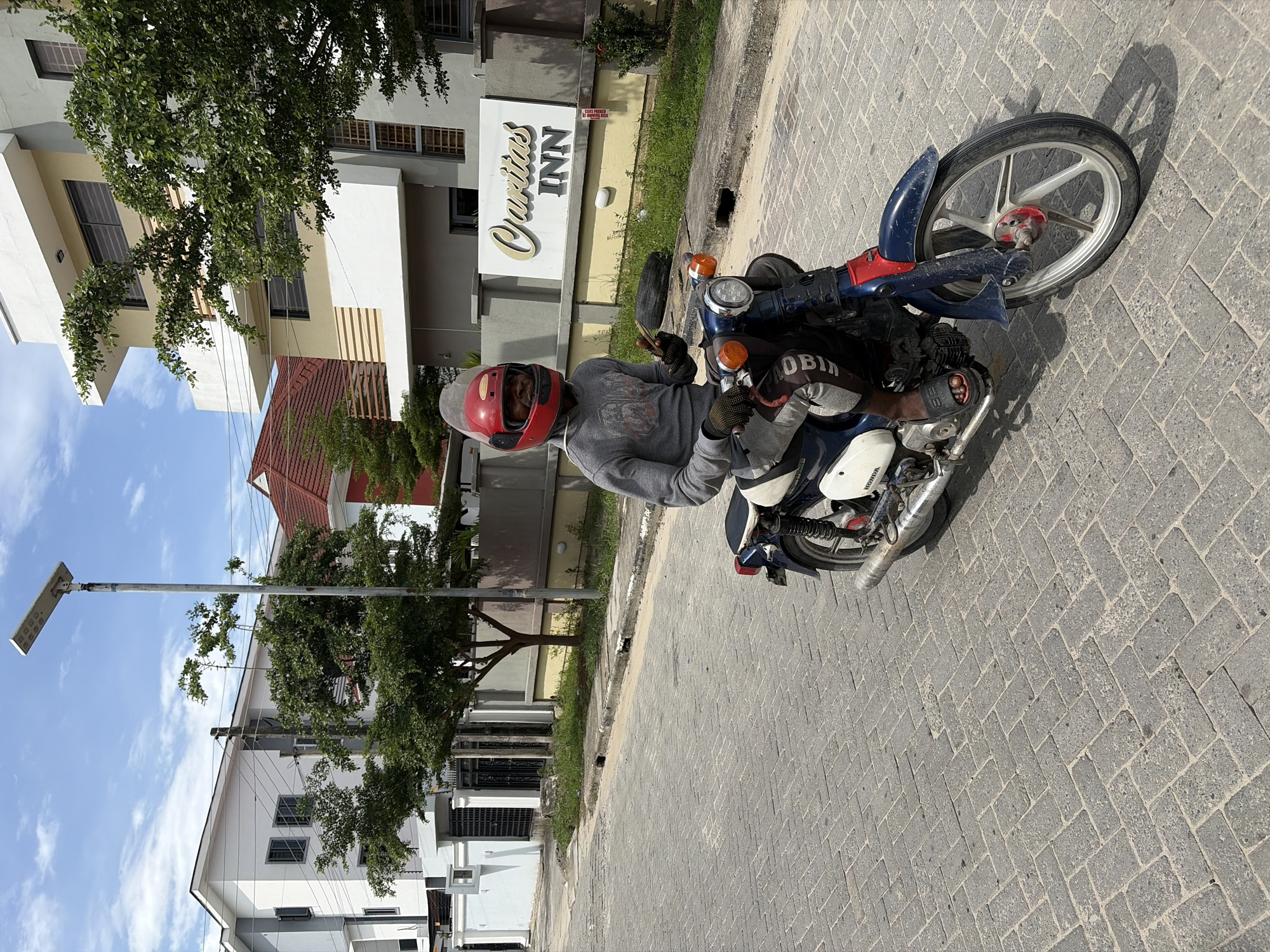 A man with dark skin wearing a red helmet, a gray sweatshirt, and gray and black pants is sitting on a blue and white Honda motorcycle on a paved road. The motorcycle is positioned near a sign that reads "Ourlins Inn." The location is Igboefon, Nigeria. A streetlight and several buildings are visible in the background.