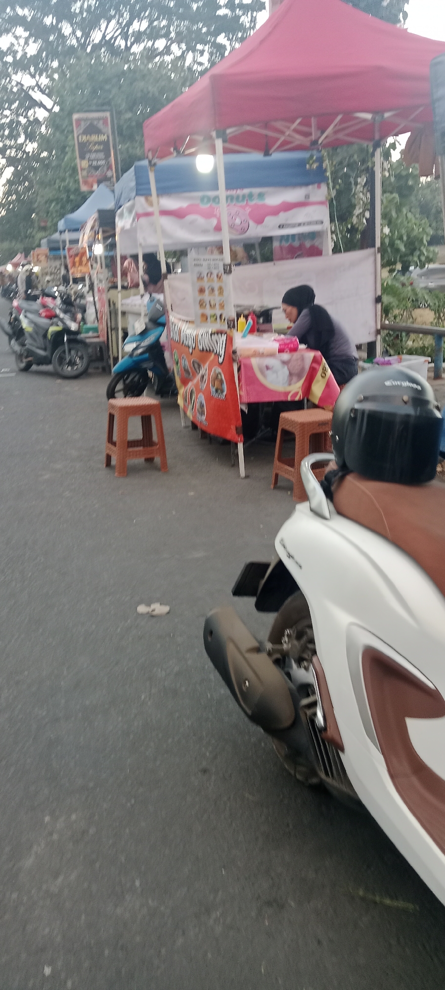 In Ngemplak, Indonesia, a street scene depicts an outdoor food stall area. A woman wearing a dark hijab and a grey shirt is seated behind a table, engaged in an activity that appears to be serving food or handling transactions. The stall is adorned with a banner featuring colorful images, possibly advertising food items like donuts. A red canopy extends over the stall, providing shade. Various motorcycles are parked nearby, suggesting this is a common mode of transport for customers. A white scooter with a brown seat is prominently positioned in the foreground, partially obscuring other elements. Several small plastic stools are placed around the stall, indicating seating for patrons. The ground surface is asphalt, typical of a street. The overall atmosphere suggests a bustling, casual marketplace setting during daylight hours.