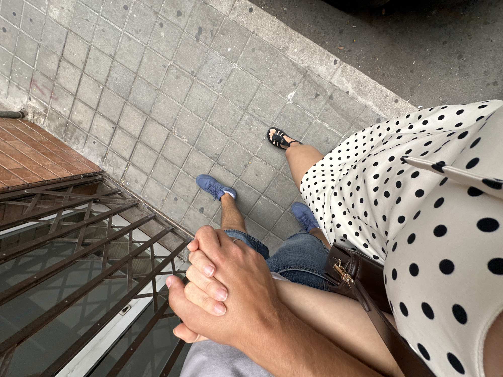 A high-angle medium shot shows a couple holding hands while walking on a city sidewalk. The individuals are identified by their attire: one is wearing blue jeans and blue sneakers, while the other is wearing a white dress with black polka dots and black sandals. Their hands are clasped in the center of the frame. The background consists of a tiled sidewalk and a brick building with a metal gate. The lighting suggests it is daytime.