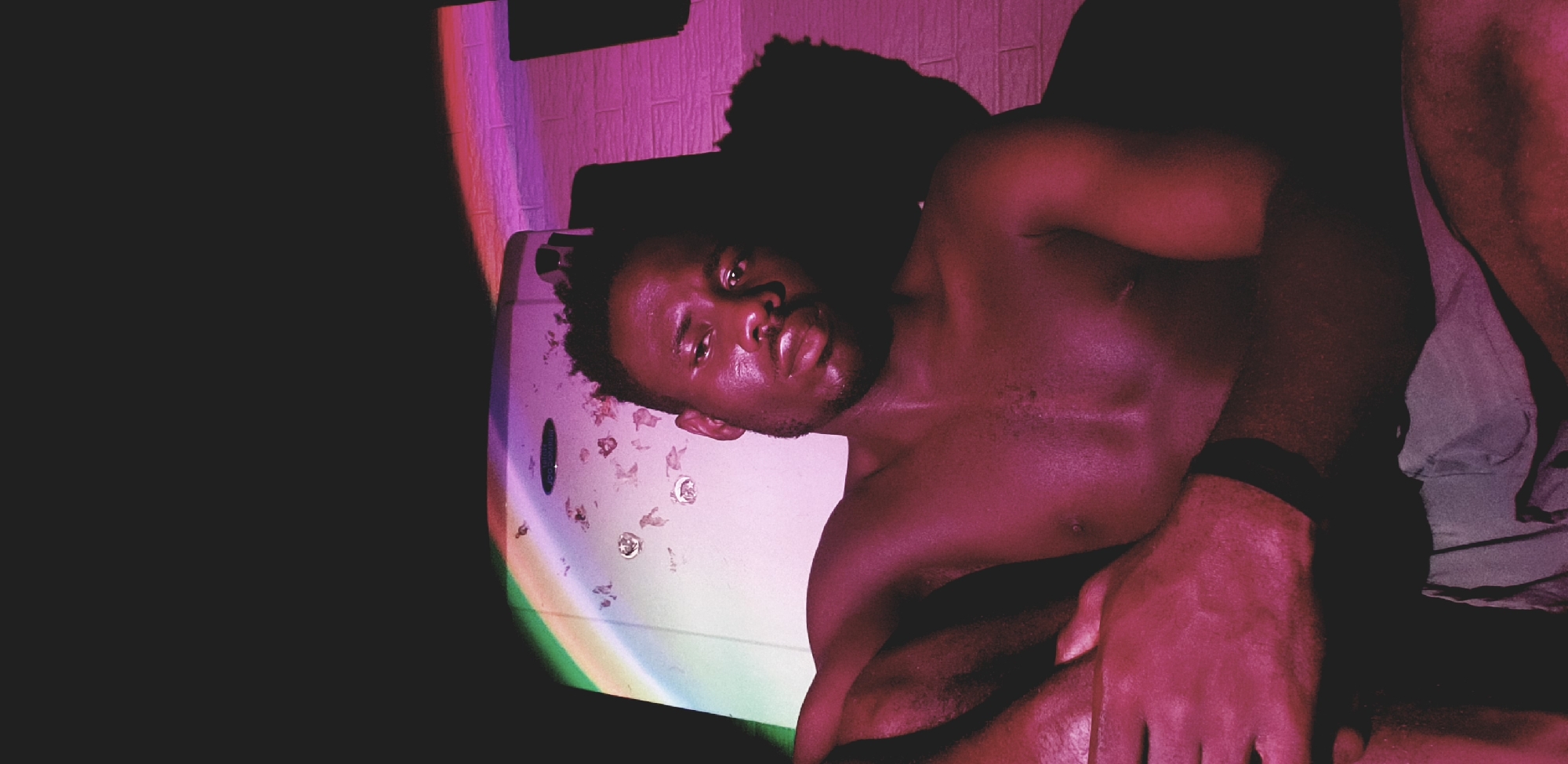 A shirtless Black man is lying down, looking at the camera. He appears to be lying next to a white jacuzzi tub with scattered petals or similar debris. There is a dark area to the left. In the background, there is a tiled wall and what appears to be a dark-colored object or shadow. A vibrant rainbow light effect shines on the side of the tub. Based on the prompt, this is in Narayi, Nigeria.