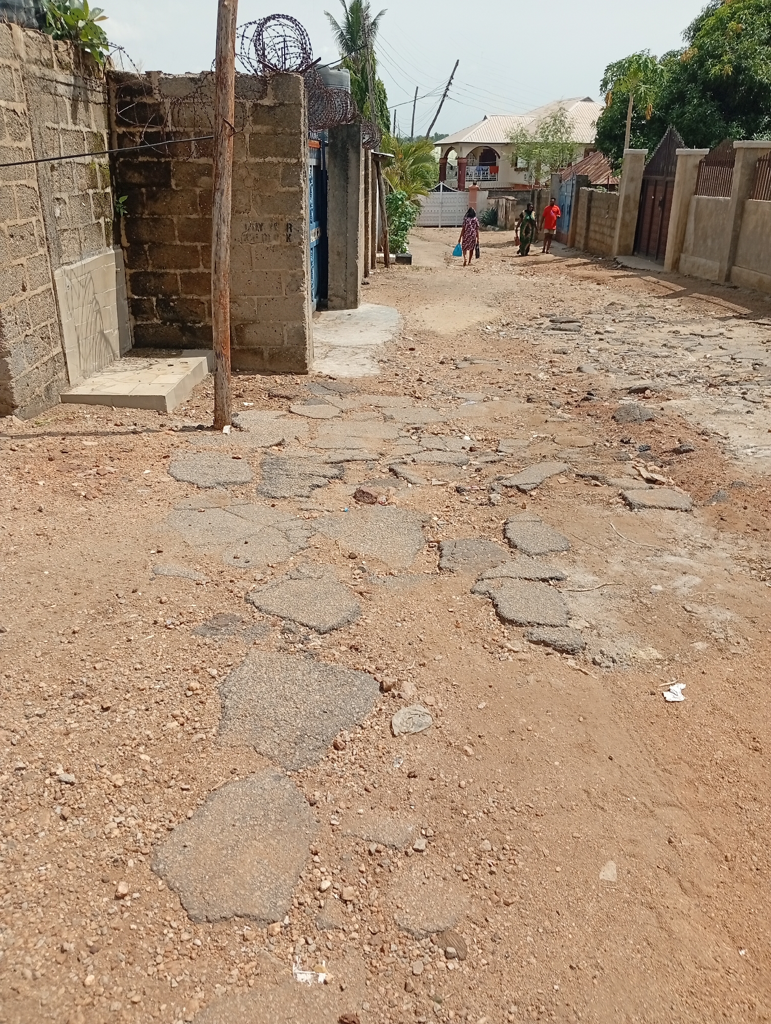 The image depicts a poorly maintained road in Adankolo, Nigeria. The road surface is severely degraded, with large sections of broken asphalt surrounded by loose gravel and dirt. A deteriorating cinder block wall topped with barbed wire stands on the left side of the road. A wooden utility pole is positioned near the wall. Three people are walking in the distance on the right side of the road. Buildings and fences are visible on either side of the road further in the background. The sky is overcast.