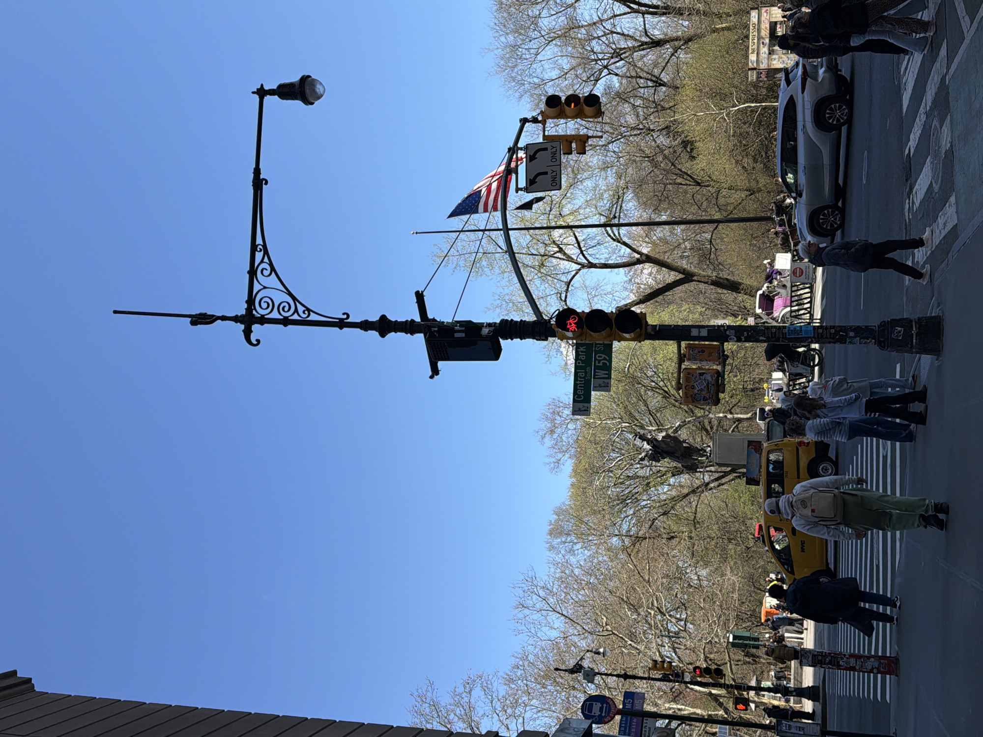 The image depicts a street scene in New York City, United States of America, specifically at the intersection of Central Park South (W 59th Street). The view is from street level looking upward.
In the foreground, a decorative lamppost and a traffic signal stand prominently. An American flag is attached to the traffic signal pole. A street sign indicates "Central Park South W 59 St". The traffic signal shows a red light for bicycles.
In the background, there is a glimpse of Central Park with bare trees. Pedestrians are visible crossing the street, and yellow taxicabs can be seen among other vehicles. A white vehicle is parked or stopped on the right side of the frame. Buildings are partially visible at the bottom and top right corners. The sky is clear and blue.