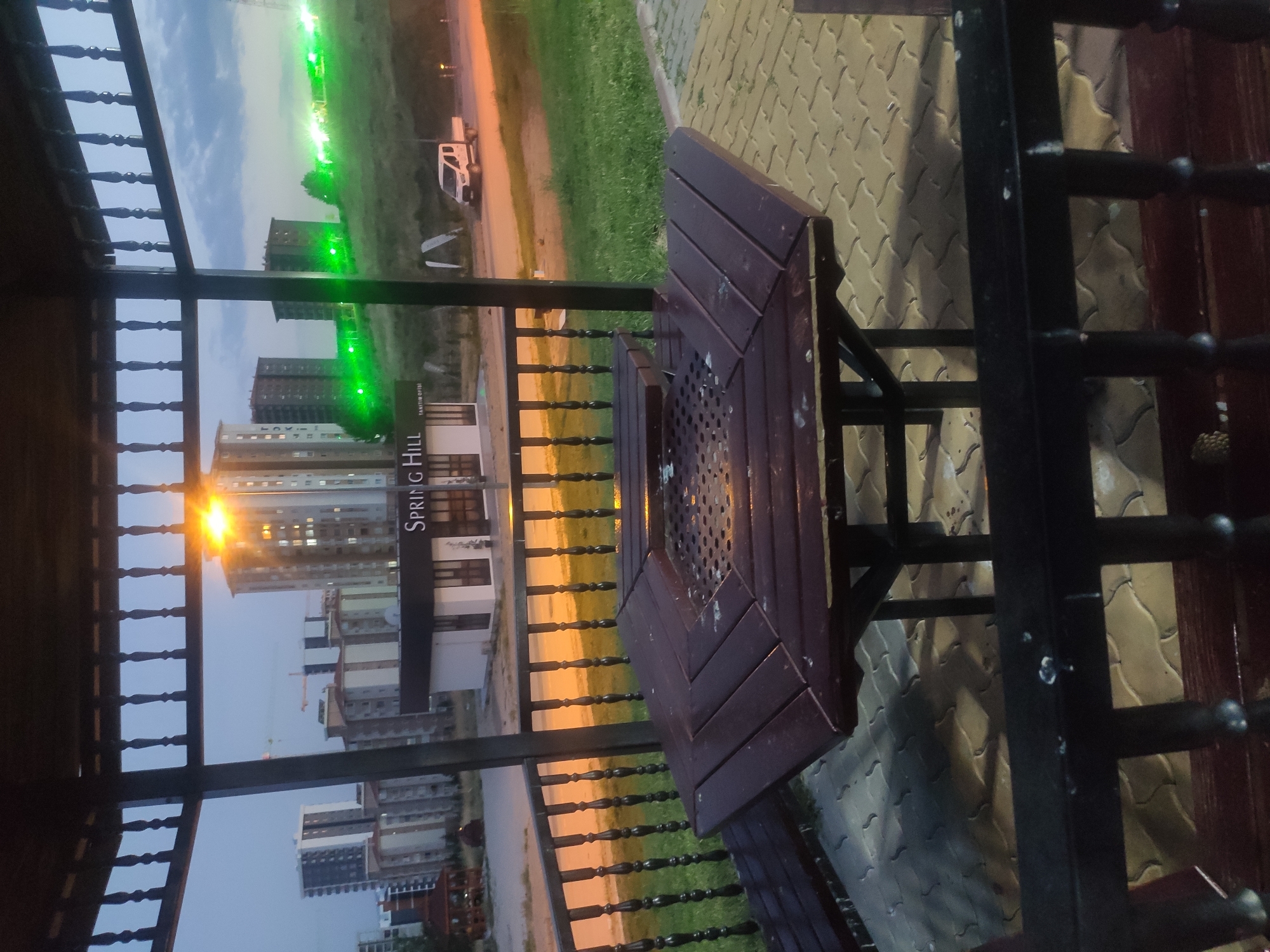 The image shows a wooden gazebo with a hexagonal table and benches. In the background, there are several apartment buildings, some of which appear to be under construction. There are also green lights on a grassy hill in the distance. A white pickup truck is parked on the street. The scene is located in Etimesgut, Turkey, and it appears to be dusk.