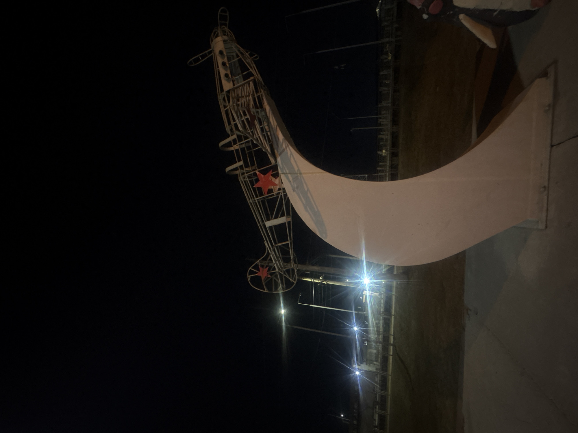 The image shows a monument in Krasnoyarsk, Russia, at night. The monument features a skeletal representation of an airplane, complete with a red star emblem. The airplane structure is juxtaposed with a curved, solid white or light-colored segment that forms part of the overall design. The scene is illuminated by streetlights, with a dark sky in the background. To the right, portions of what appears to be a concrete walkway and adjacent structures are visible.
