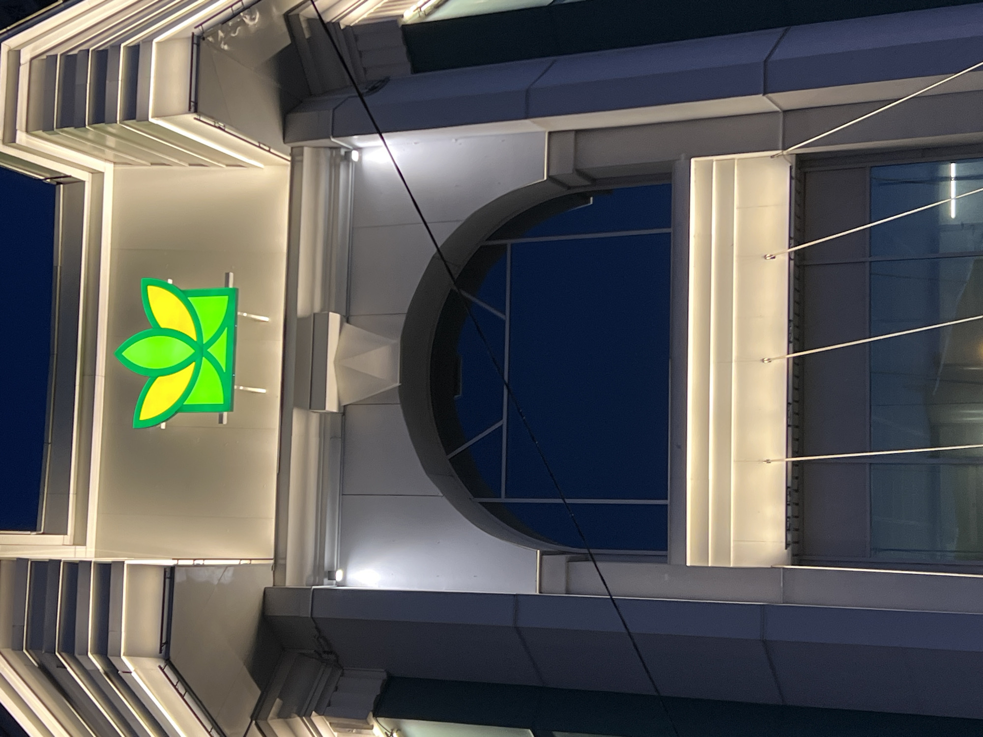 The image shows a facade of a building at night, likely in an urban setting. The building features a prominent, illuminated green and yellow logo on its upper section, possibly representing a company or organization. Below the logo, a large arched window with a dark blue interior is visible. The building's architecture includes modern design elements with sharp lines and embedded lighting. Electrical wires are visible crossing the frame in front of the window and building. The overall impression is of a commercial or institutional building with a contemporary aesthetic, illuminated for nighttime visibility. The city is Unknown, Russia.