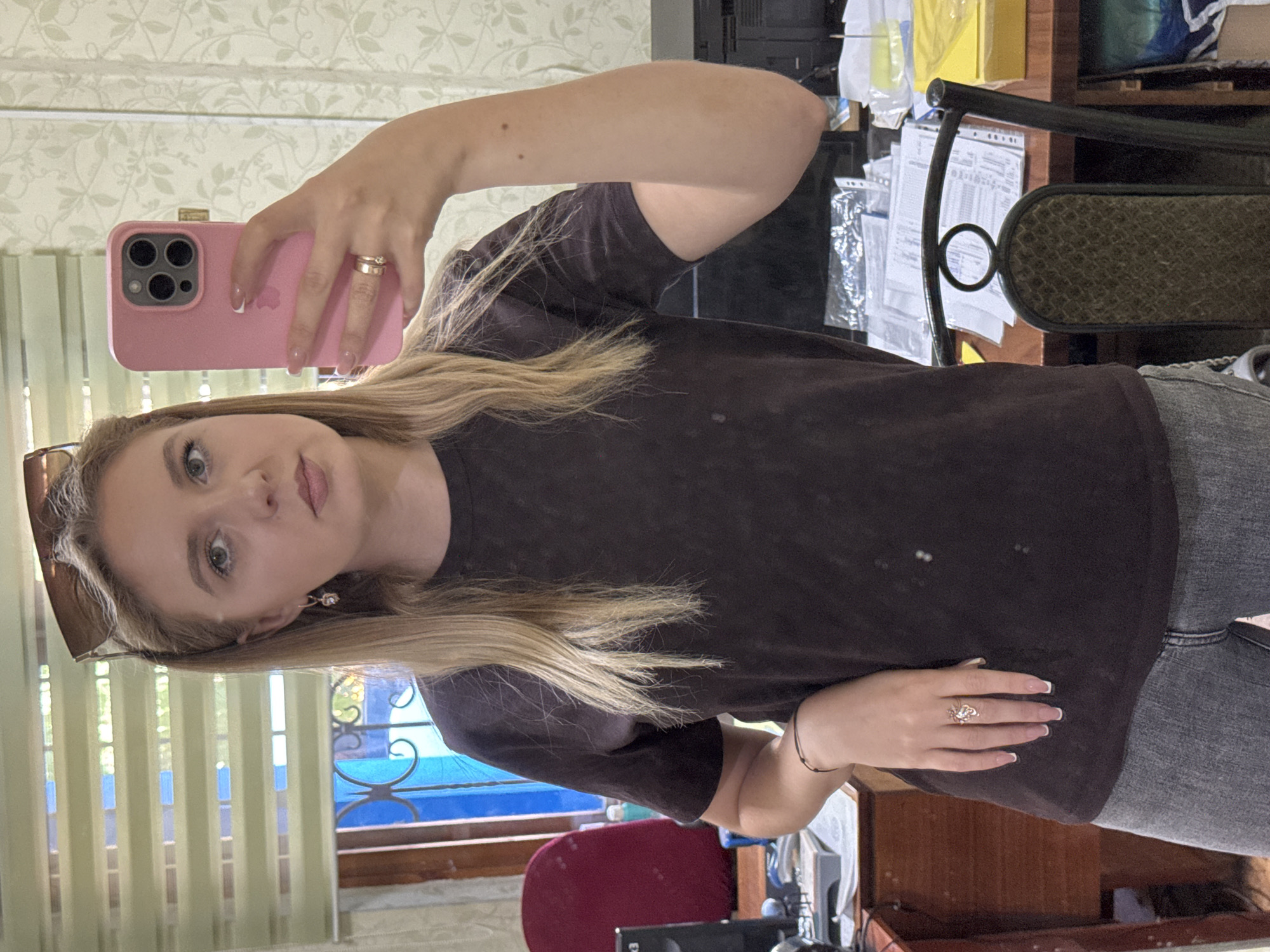 A young woman with blonde hair and blue eyes is taking a selfie in an office environment. She is wearing a dark t-shirt, grey jeans, a ring on her right ring finger, a bracelet on her left wrist, and earrings. Her sunglasses are resting on top of her head. The woman is holding a pink smartphone in her right hand. In the background, there is a desk with papers and a computer monitor, a chair, and a window with blinds. The lighting suggests it is daytime.