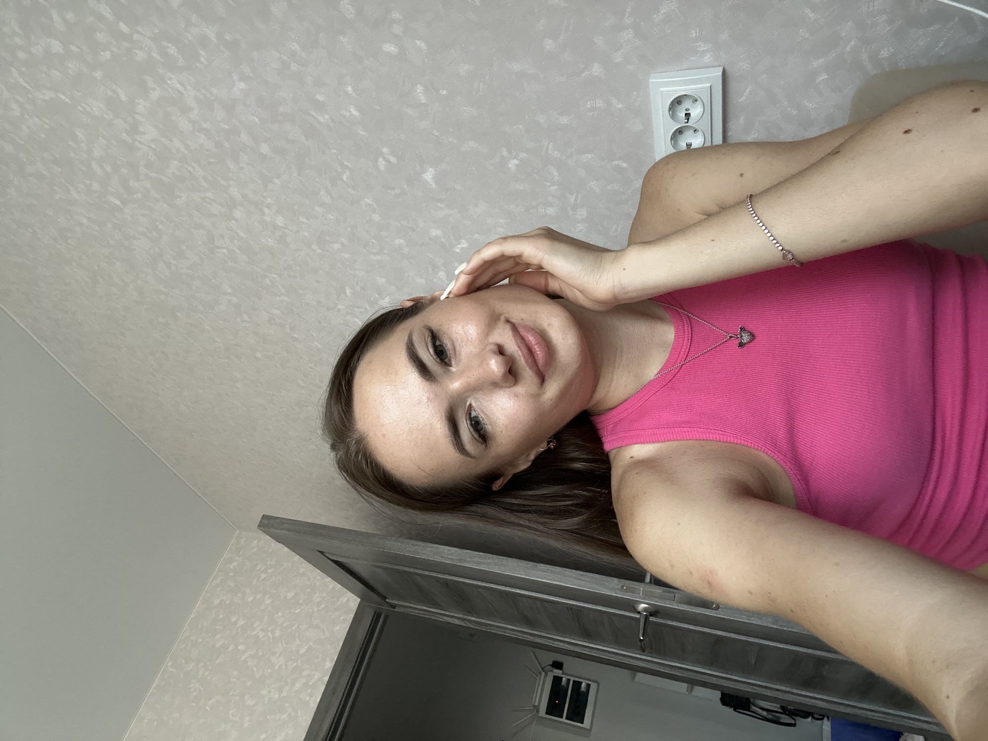 The image shows a young woman with light skin and brown hair. She is wearing a bright pink tank top and a silver necklace with a pendant. Her left hand is resting on her cheek, and she is looking at the camera with a slight smile. The background appears to be a room with textured wallpaper and a closed door. There is also a white electrical outlet visible on the wall. The lighting is soft and natural. The image is a close-up shot, focusing on the woman's face and upper body.