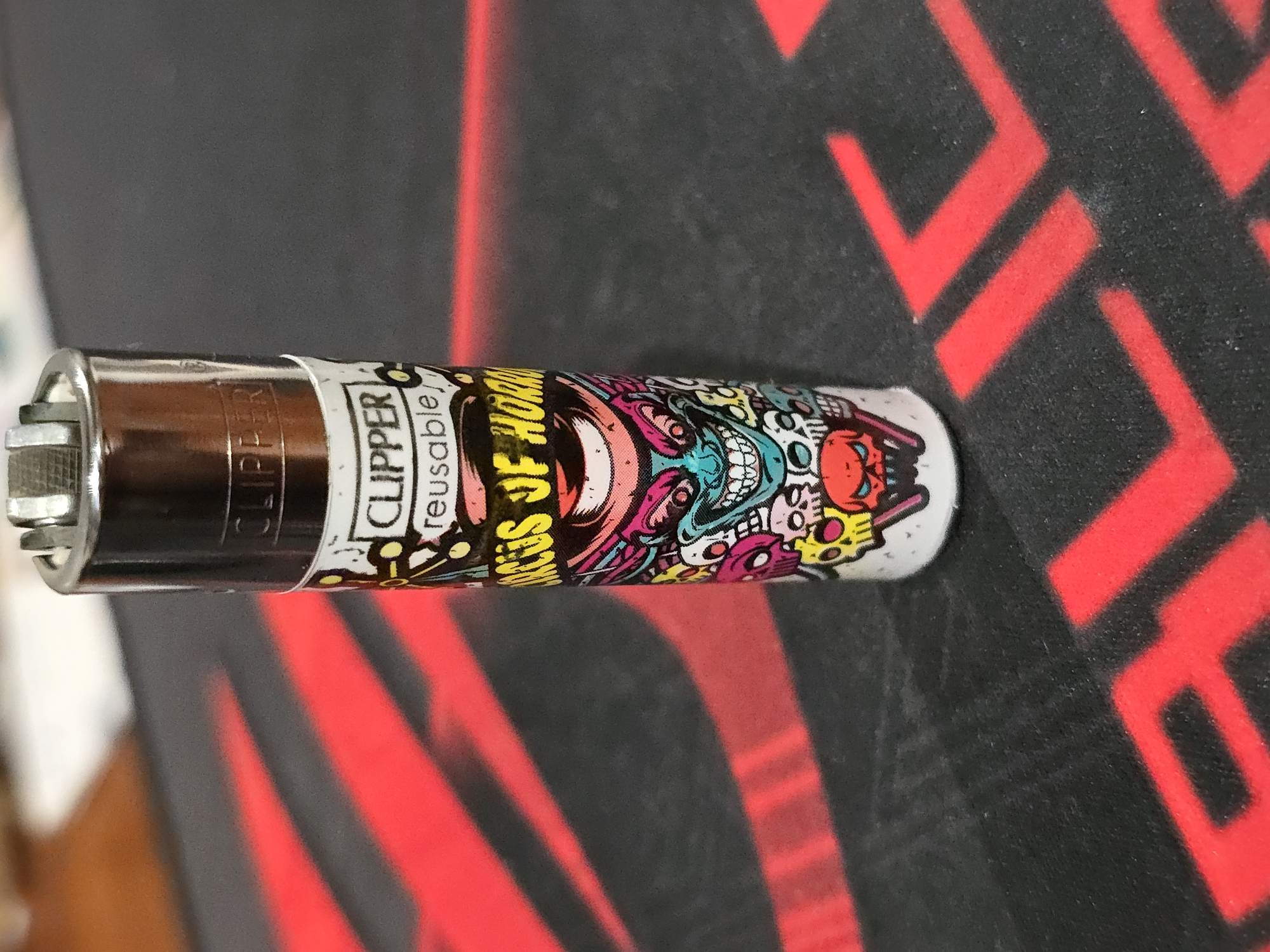 A close-up, eye-level shot shows a Clipper brand lighter with a "Circus of Horror" design. The lighter is silver on top with a red wheel, and the body is white with a colorful, cartoonish graphic of various monstrous faces and skulls. The graphic features bright pink, blue, yellow, and red elements. The lighter stands upright on a black surface with red abstract shapes or logos. The background is out of focus and appears to be a wooden table or countertop. The lighting is even, highlighting the details of the lighter and its design. There is no discernible indication of location within a city, nor any people present.