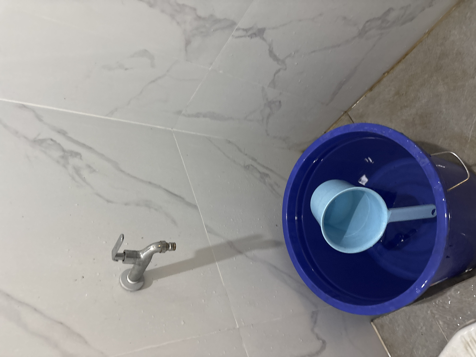 A silver faucet is attached to a white tiled wall with subtle gray marble patterns. Below the faucet, a large blue plastic bucket contains water and a light blue plastic ladle. The floor is tiled with gray square tiles. The image appears to be taken in a bathroom or utility area. This scene is located in the city of Bekasi, Indonesia.