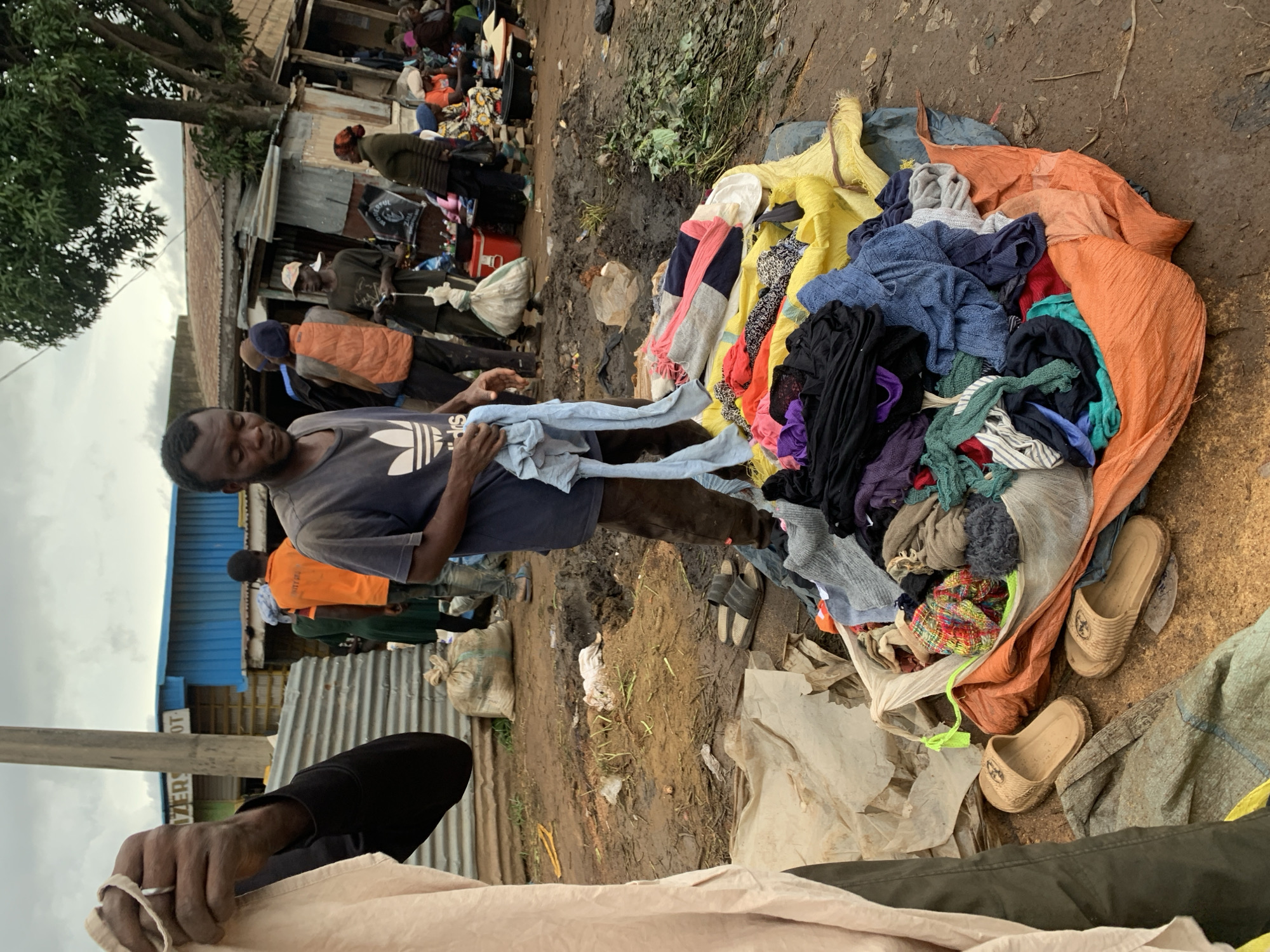 In Gura Top, Nigeria, a bustling market scene unfolds on a cloudy day. The ground, a mixture of mud and dirt, is strewn with various items, suggesting a chaotic yet vibrant commercial environment. In the foreground, a man in a dark t-shirt with an Adidas logo holds a piece of light-colored fabric, his left hand wearing a ring. He appears to be sorting through or displaying clothing, which is piled high on a yellow and orange tarp. The pile consists of numerous garments in various colors and textures, including sweaters, t-shirts, and patterned fabrics. Two pairs of light brown sandals are placed near the pile of clothes. In the background, several individuals are engaged in market activities. A man in an orange vest stands near other people, some of whom are obscured by the foreground subjects. Further back, a row of market stalls with corrugated metal roofing and blue siding indicates the commercial infrastructure of the area. Bags, possibly for carrying goods, are visible, along with other assorted items that contribute to the overall impression of a lively marketplace.