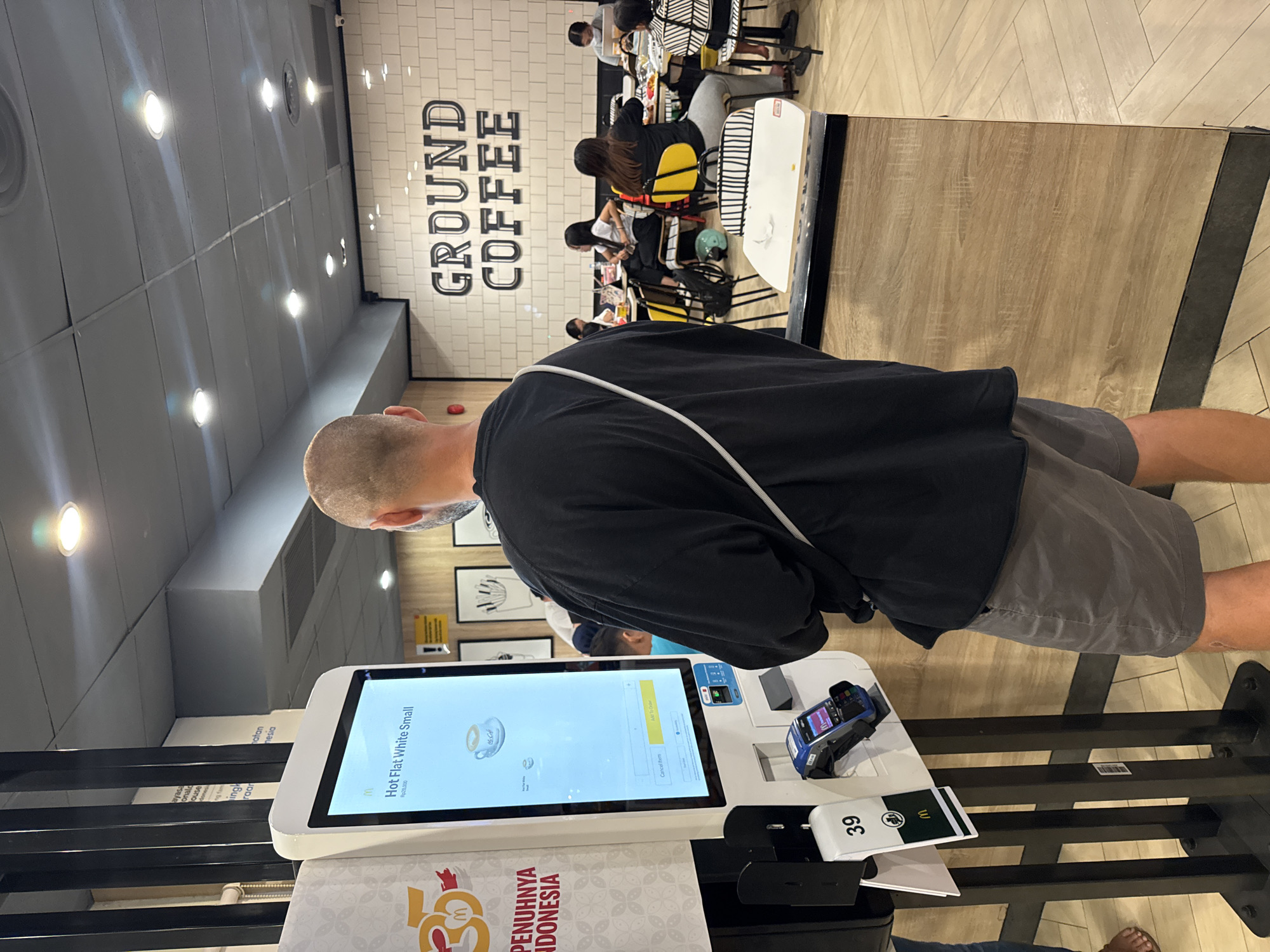 The image shows a person ordering at a self-service kiosk in a McDonald's restaurant. The screen displays a "Hot Flat White Small" beverage choice, priced at Rp28,000. Below the screen is the text "PENUHNYA INDONESIA". A card reader and a numbered order ticket (39) are visible on the counter. Behind the person are other diners at tables, and a wall with the words "GROUND COFFEE". The image was taken in Kuta Selatan, Indonesia.