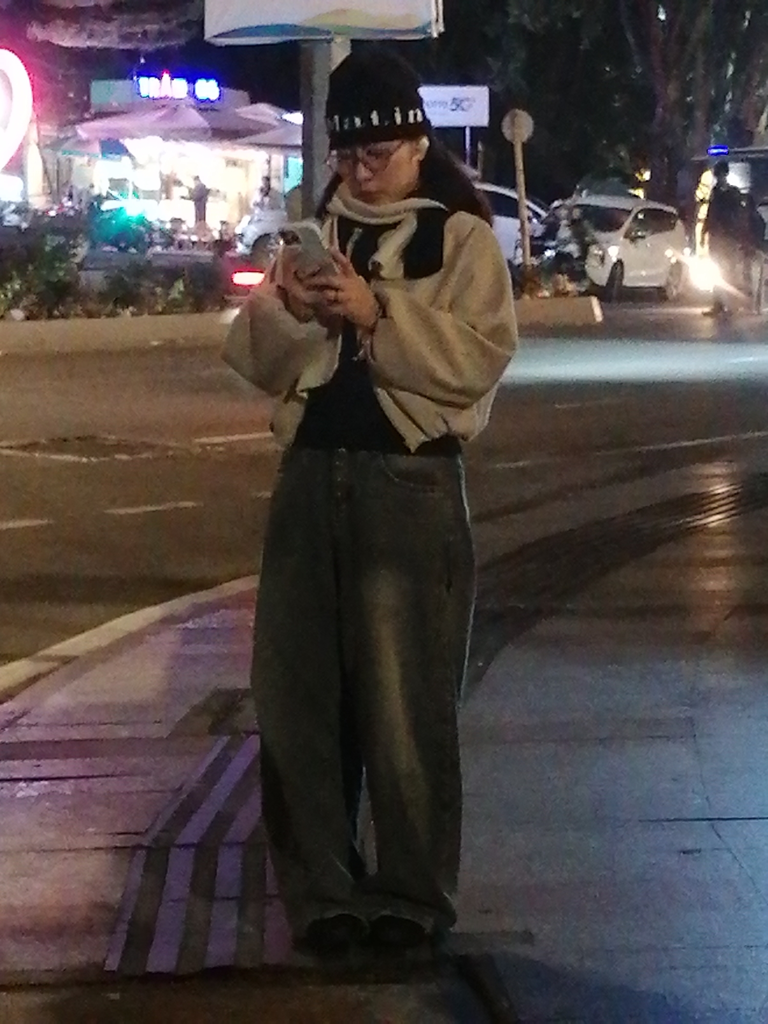 In Da Lat, Vietnam, a person stands on a sidewalk next to a street at night. The individual is wearing a black beanie, glasses, a light-colored jacket over a dark shirt, and loose-fitting dark pants. They are holding a smartphone in their hands, looking down at it. The background shows a street with cars, some of which are illuminated, and buildings with neon signs. The sidewalk has a textured surface with alternating dark and lighter colored stripes. The lighting suggests it is nighttime, with artificial lights from buildings and vehicles creating reflections on the wet street surface.