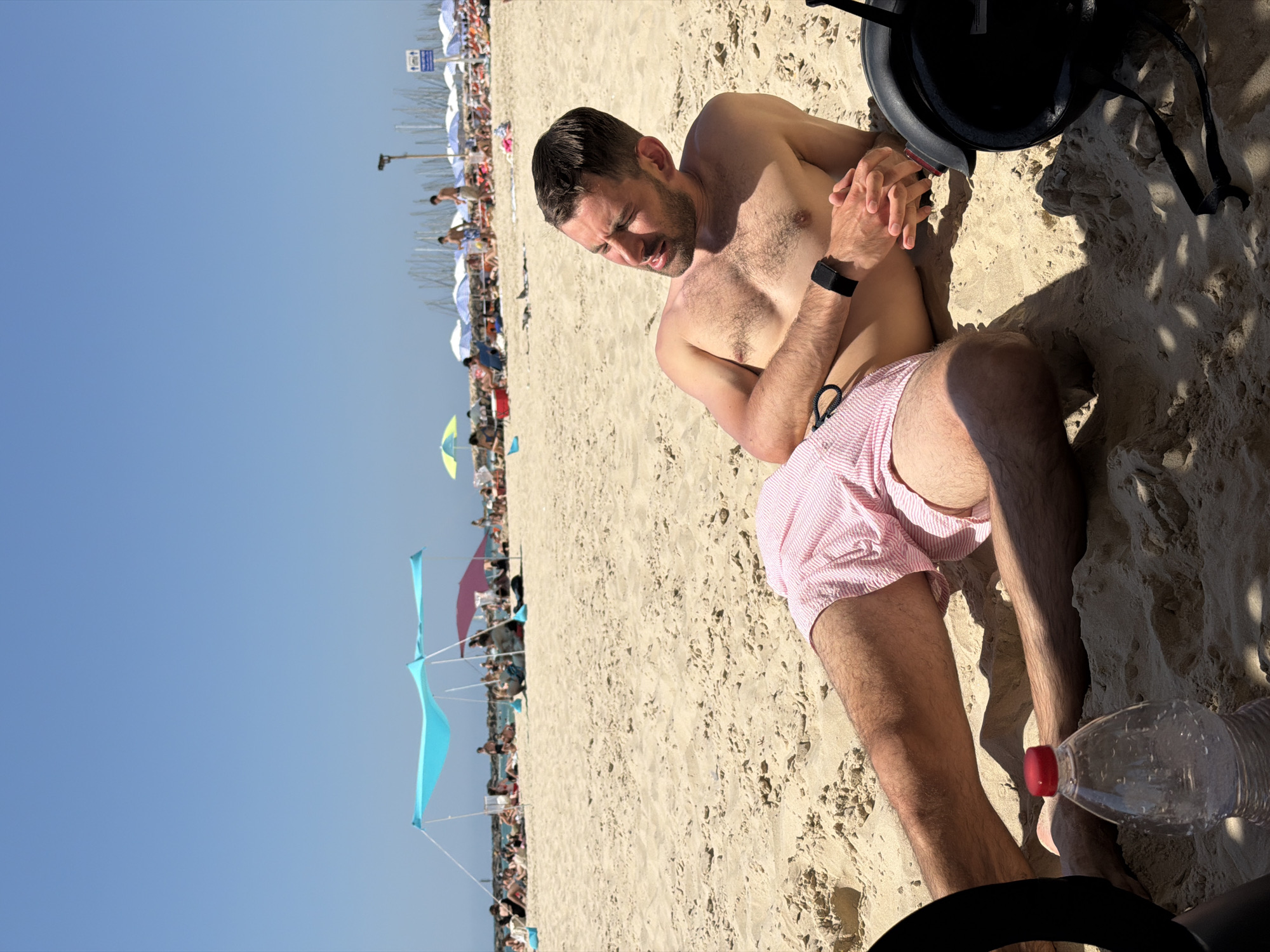 A man is reclined on a beach, possibly in Tel Aviv, Israel, given the style of umbrellas and beach atmosphere in the background. He is shirtless, wearing red and white striped shorts, and a black smartwatch. He is resting on the sand with his hands clasped together. Nearby, there is a black helmet and a partially visible plastic water bottle with a red cap. The background shows a crowded beach with various colored umbrellas, kites, and other beach-goers. The sky is clear blue.