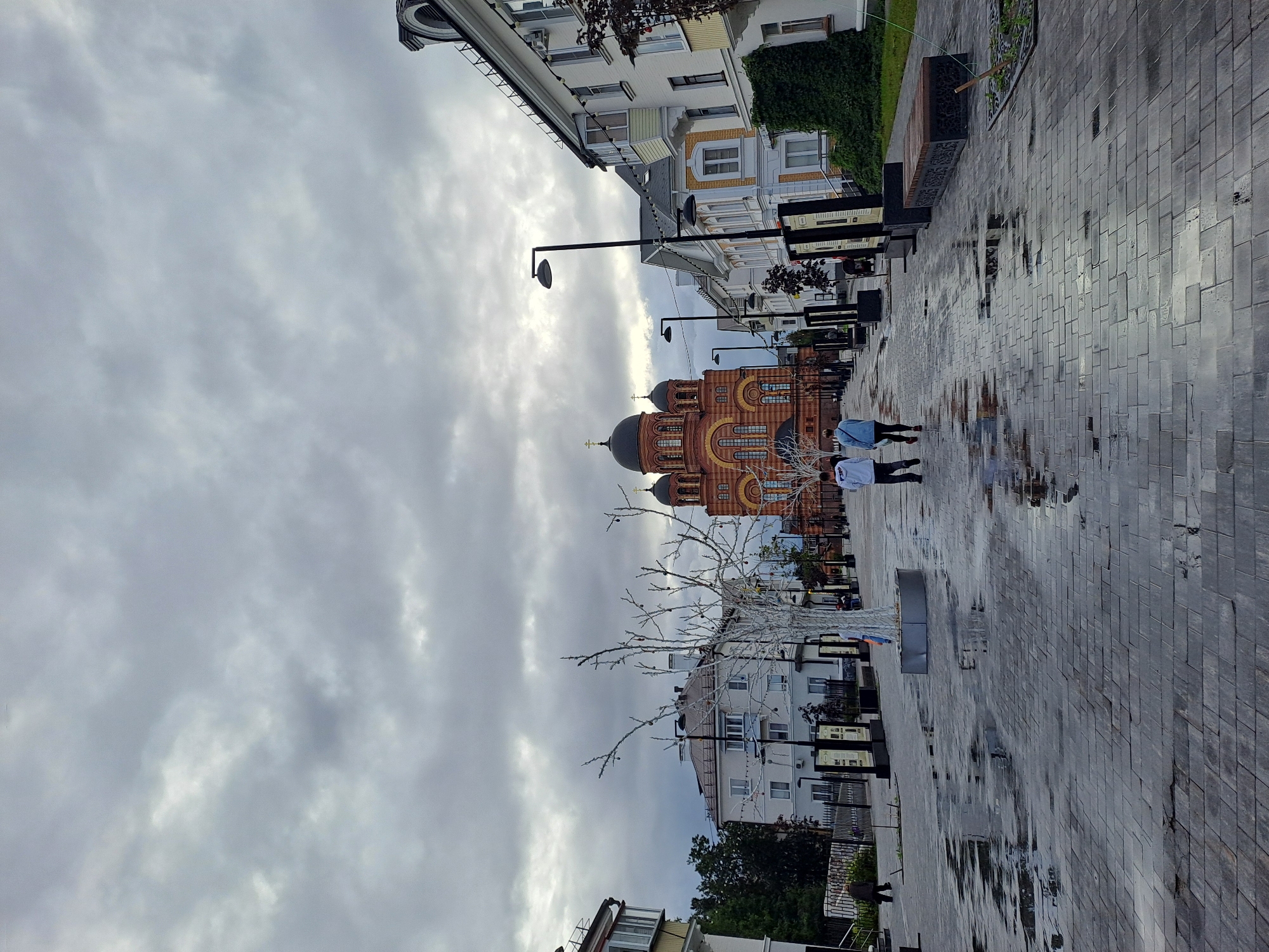 The image captures a paved pedestrian walkway in Elektrostal, Russia, leading towards a large, ornate orange and red church with multiple domes. The sky is overcast with gray clouds. The walkway is wet and reflective, indicating recent rain. Several people are walking on the path; two individuals in the foreground are walking away from the camera, dressed in casual clothing. Along the sides of the walkway, there are buildings, streetlights, and decorative white trees that appear to be illuminated. The environment suggests a cloudy day in an urban setting with a prominent religious structure.