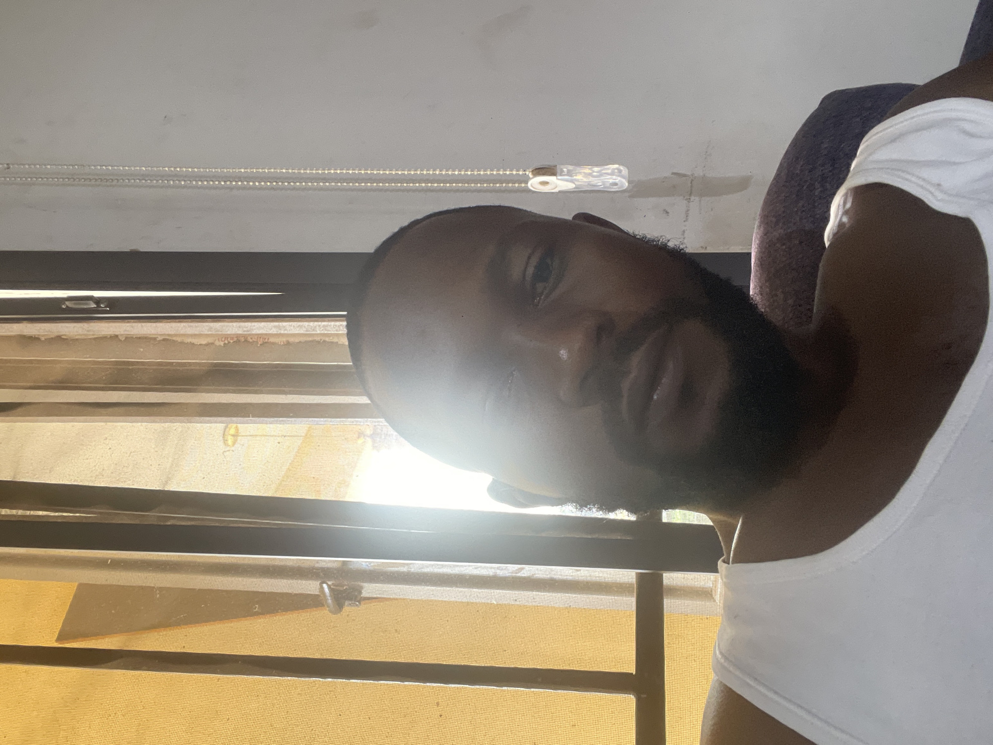 A man is indoors, wearing a white tank top. He has dark skin, short hair, and a full beard. He appears to be looking out of a window with light-colored blinds. Based solely on the visual information available, the location in Abuja, Nigeria, cannot be confirmed.