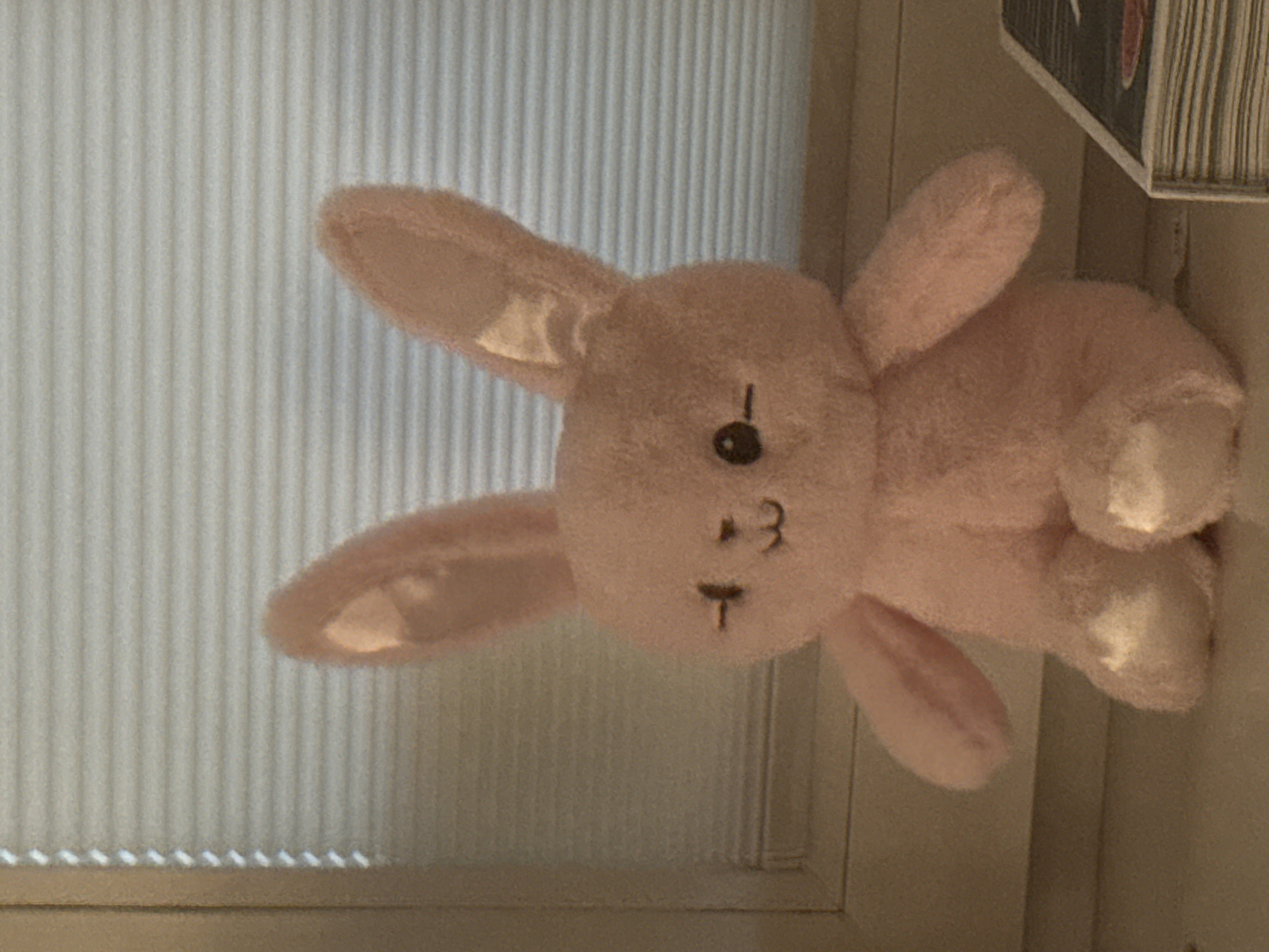 A pink stuffed bunny with long ears is sitting on a ledge in front of closed blinds. The bunny has embroidered eyes and mouth. The bunny's feet are a lighter shade of pink, and appear to be made of a different material. To the right of the bunny, the corner of a book is visible. The book has a black cover with white text and red accents. The setting appears to be indoors, with the blinds suggesting a window.