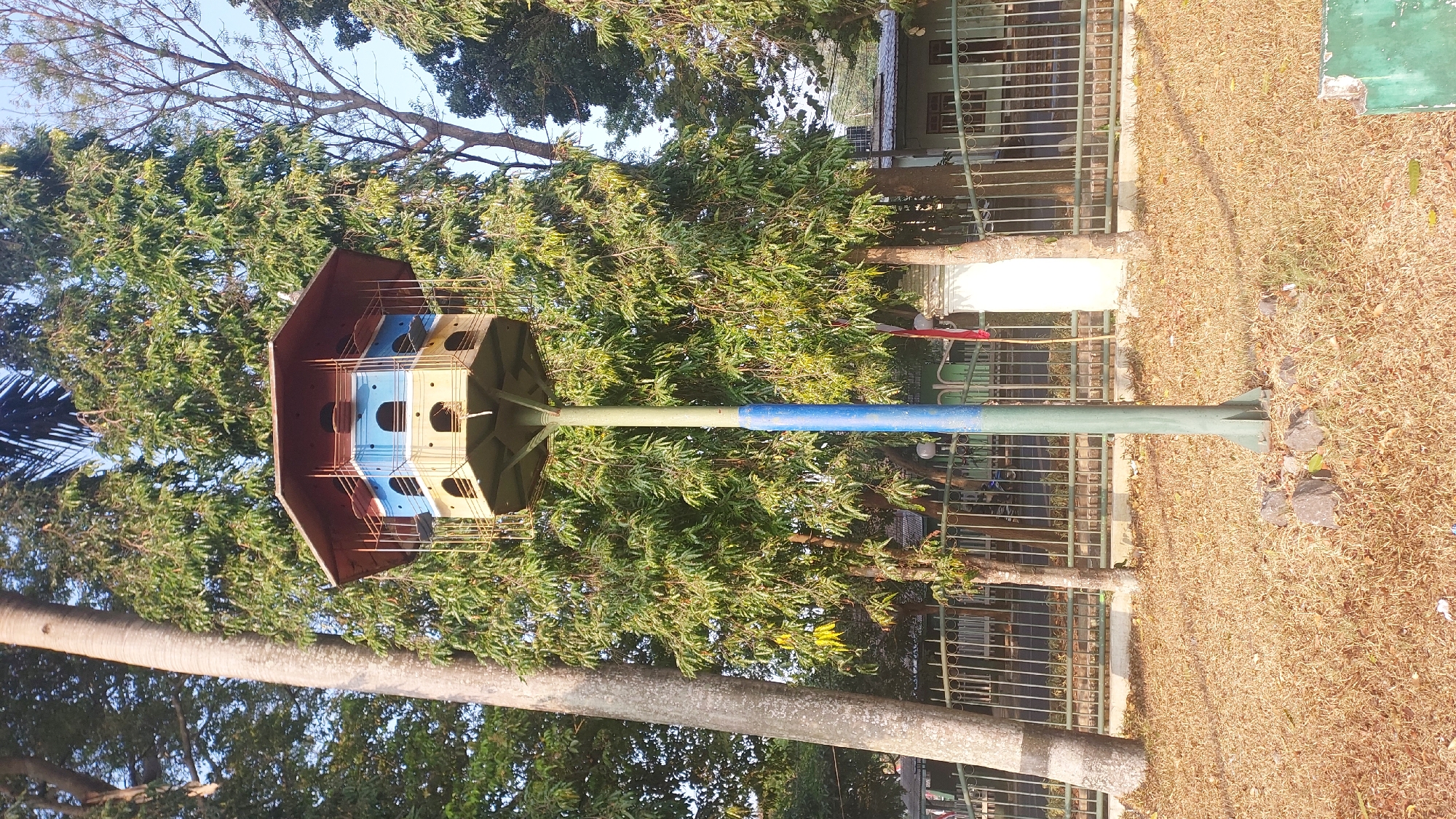 This is a medium shot, eye-level, full-color photograph of a multi-tiered birdhouse on a pole in front of a metal fence and a building. The birdhouse is octagonal with a reddish-brown roof and yellow and blue sections below. It is constructed of wood and metal, with openings for birds. The pole supporting the birdhouse is green and blue, with a metal base anchored by stones. The foreground consists of dry, brown grass and fallen leaves. Behind the fence, a building with windows is visible. Lush green trees with dense foliage dominate the background. The sky is clear and pale blue. The scene appears to be outdoors in a park-like setting, possibly in the city of Malang, Indonesia, given the foliage and architectural style. No people are present in the frame.