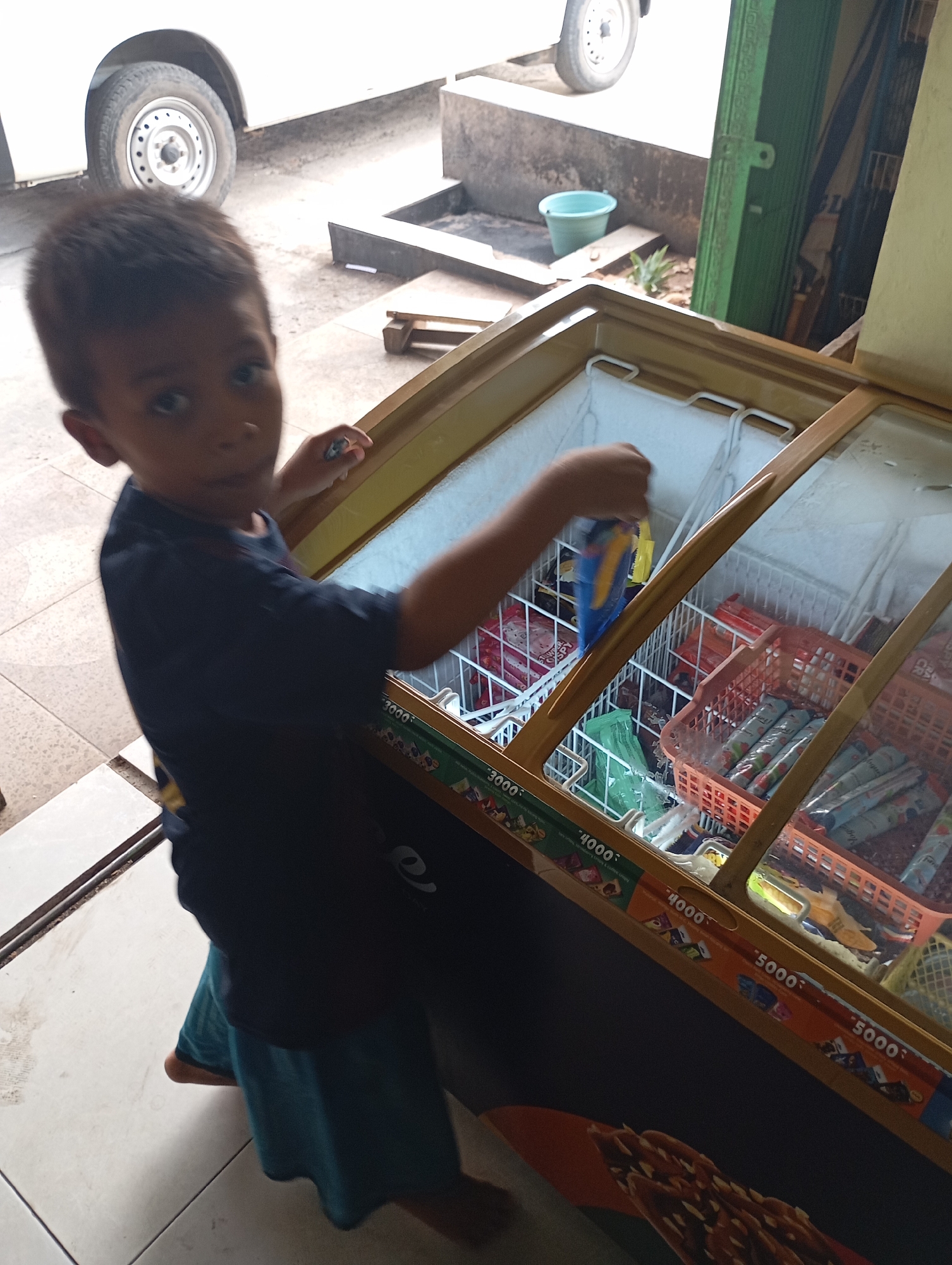 In Metro City, Indonesia, a young boy, appearing to be around 5 years old, stands in front of an open freezer filled with frozen treats. He is wearing a dark blue t-shirt and teal shorts, and his left hand is reaching into the freezer, holding a blue-wrapped popsicle. The freezer's glass lid is open, revealing rows of various frozen desserts, including some in orange plastic baskets. The freezer itself has a golden-colored frame and a decorative strip along the bottom with prices indicated by "3000", "4000", and "5000". The environment appears to be outdoors, possibly a street or sidewalk, with a tiled floor and a white vehicle parked in the background. A green trash bin is visible near a building.