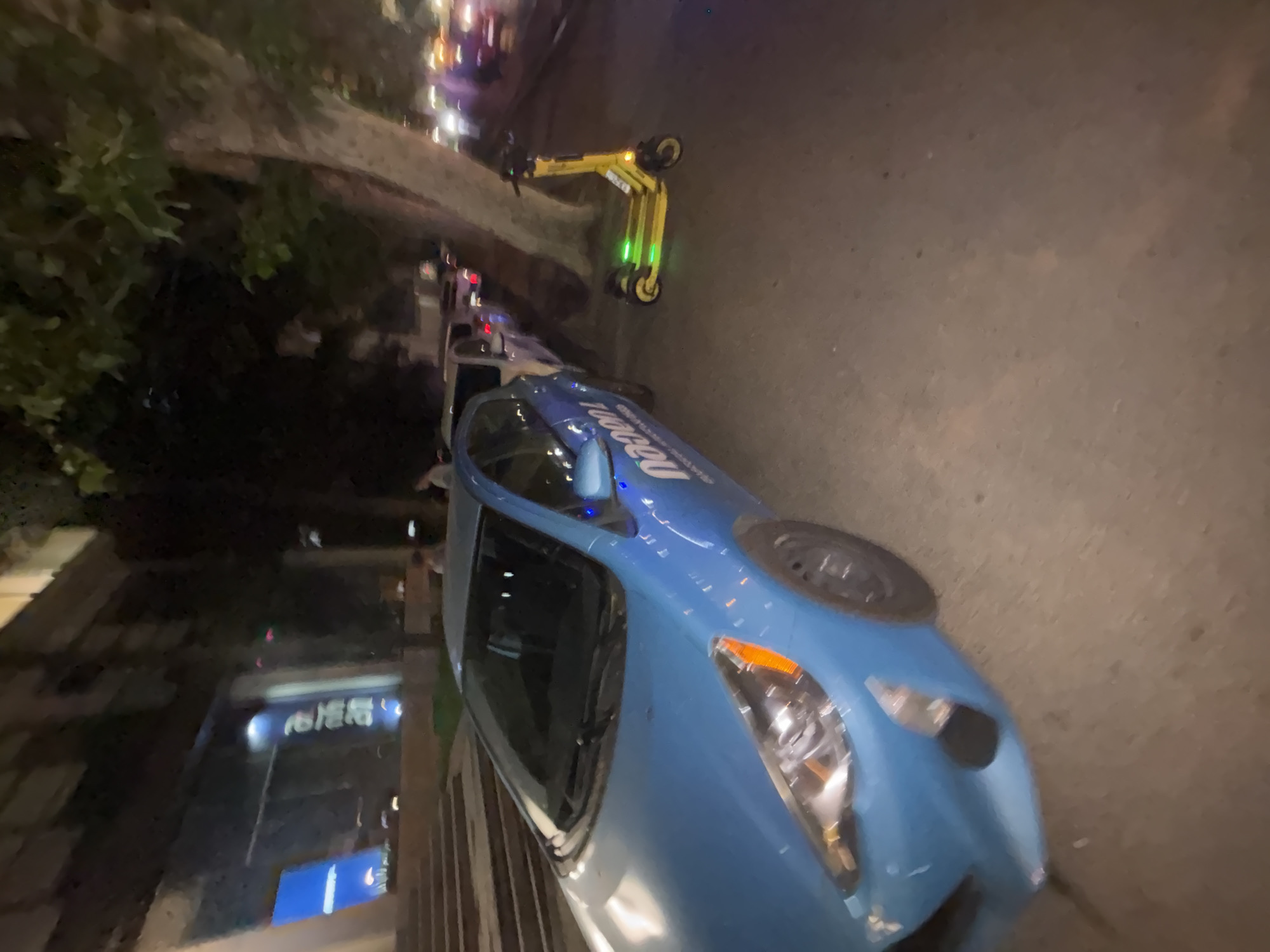 In Tbilisi, Georgia, at night, a blue car with a "Nano" logo is parked on a street. Next to the car, a yellow electric scooter with green lights is parked. Buildings and trees line the street, and there are several people in the background. The scene appears to be a busy urban area.