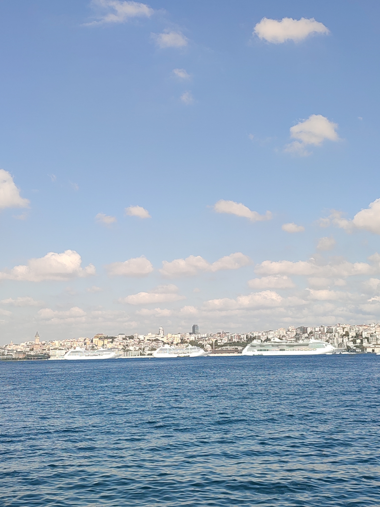 A wide shot captures the city of Üsküdar, Turkey, from across the water, likely the Bosphorus Strait. The foreground is dominated by the deep blue, slightly rippled water of the strait. In the midground, a line of large white cruise ships is docked along the Üsküdar waterfront, their sleek, modern structures contrasting with the older cityscape behind them.
The Üsküdar skyline is a dense collection of buildings, varying in height and architectural style, rising up from the waterfront. Prominent structures include a distinctive tall, dark tower on the left side of the skyline, likely the Galata Tower, and other modern buildings. The city stretches horizontally across the frame.
The background is a bright blue sky with scattered, fluffy white clouds. The lighting suggests it is daytime, possibly late morning or early afternoon, with clear visibility. There are no people visible in the frame. The overall impression is a serene, picturesque view of a vibrant coastal city.
