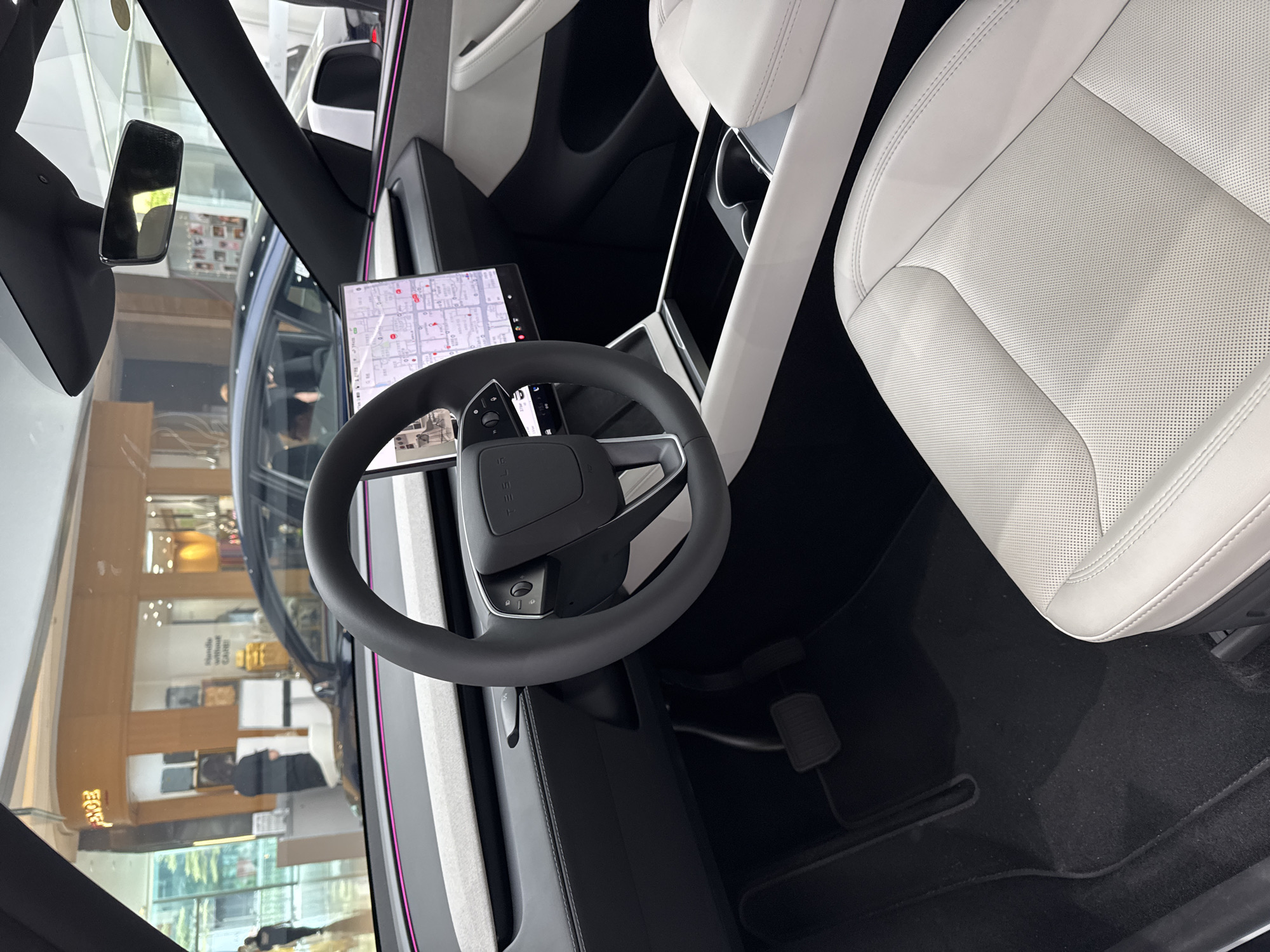 Here is a description of the image:
The image shows the interior of a car, likely a Tesla, focusing on the driver's area. A steering wheel is prominently displayed in the center, with a digital display screen visible above it, showing a map. To the right is a white leather seat. The car appears to be parked indoors, with a view through the windshield revealing a commercial space, possibly a shopping mall. The location is likely in the Tianhe District of China, given its modern appearance. Inside the car, the color scheme is primarily black and white, giving it a sleek and modern look. There are no people visible inside the car, only reflections through the windshield. The environment appears to be well-lit.