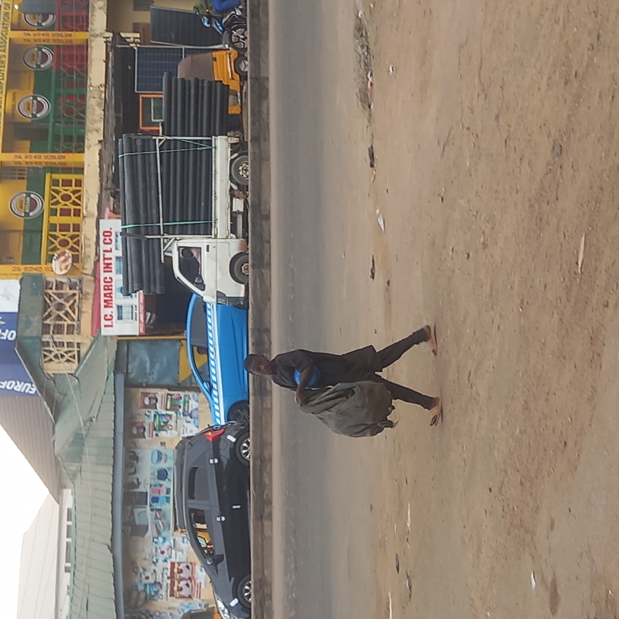 A man carries a sack along a road in Dadi Riba, Nigeria. The road is lined with parked cars and a truck loaded with black pipes. Businesses are visible behind the vehicles. The ground is dirt.