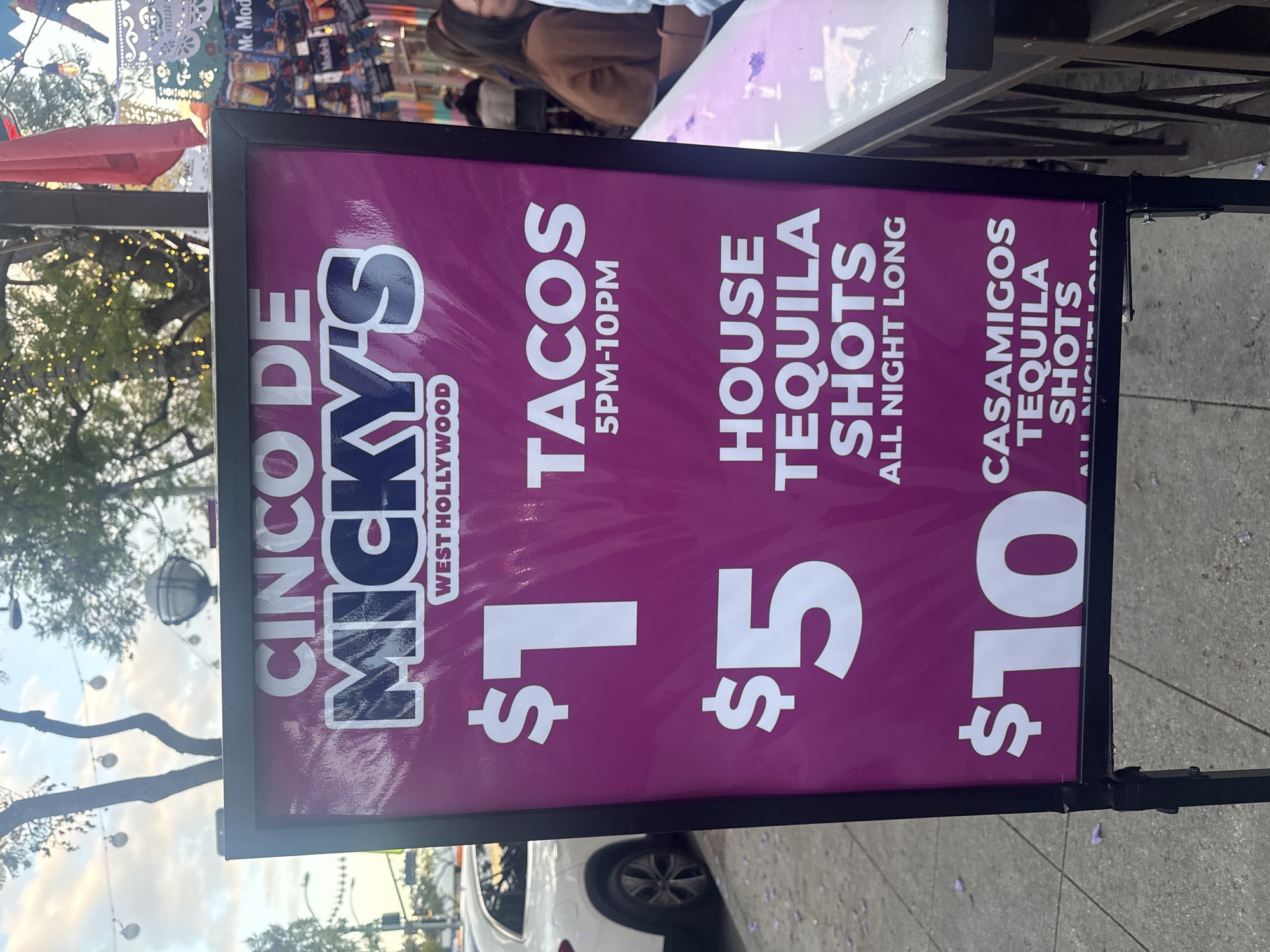 Here is a detailed description of the image:
The image shows a promotional sign for Cinco De Micky's in West Hollywood. The sign is purple and features the following deals: $1 tacos from 5PM-10PM, $5 house tequila shots all night long, and $10 Casamigos tequila shots. The sign is positioned on a city street. A small section of a white car can be seen parked at the bottom edge of the sign. A group of people are shown at the top edge of the sign. Other establishments with advertising signs are slightly visible in the top left corner.