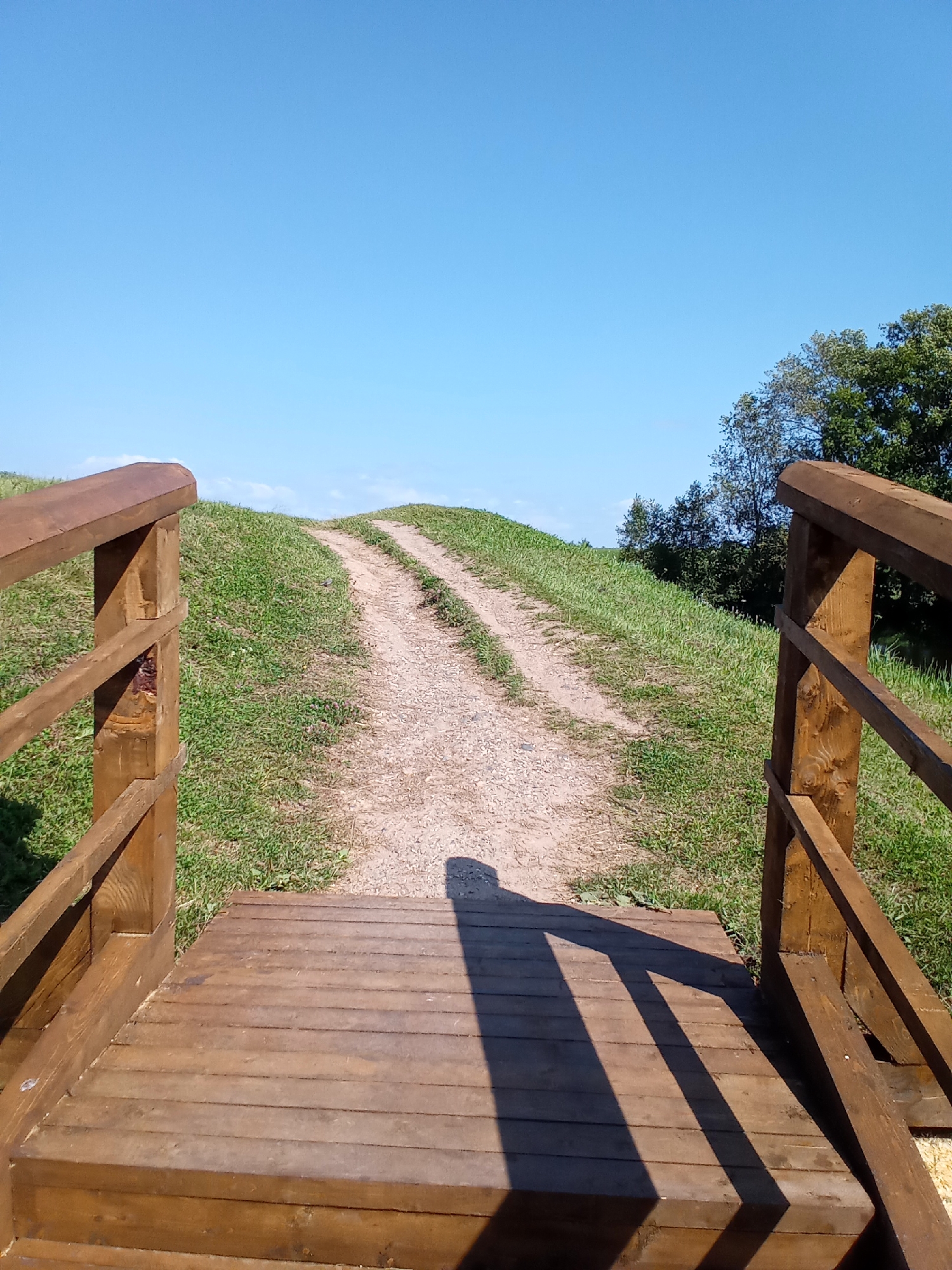 The image depicts a wooden walkway and railing leading to a dirt path that ascends a grassy hill under a clear blue sky. Trees are visible in the background on the right. The scene appears to be an outdoor recreational area.