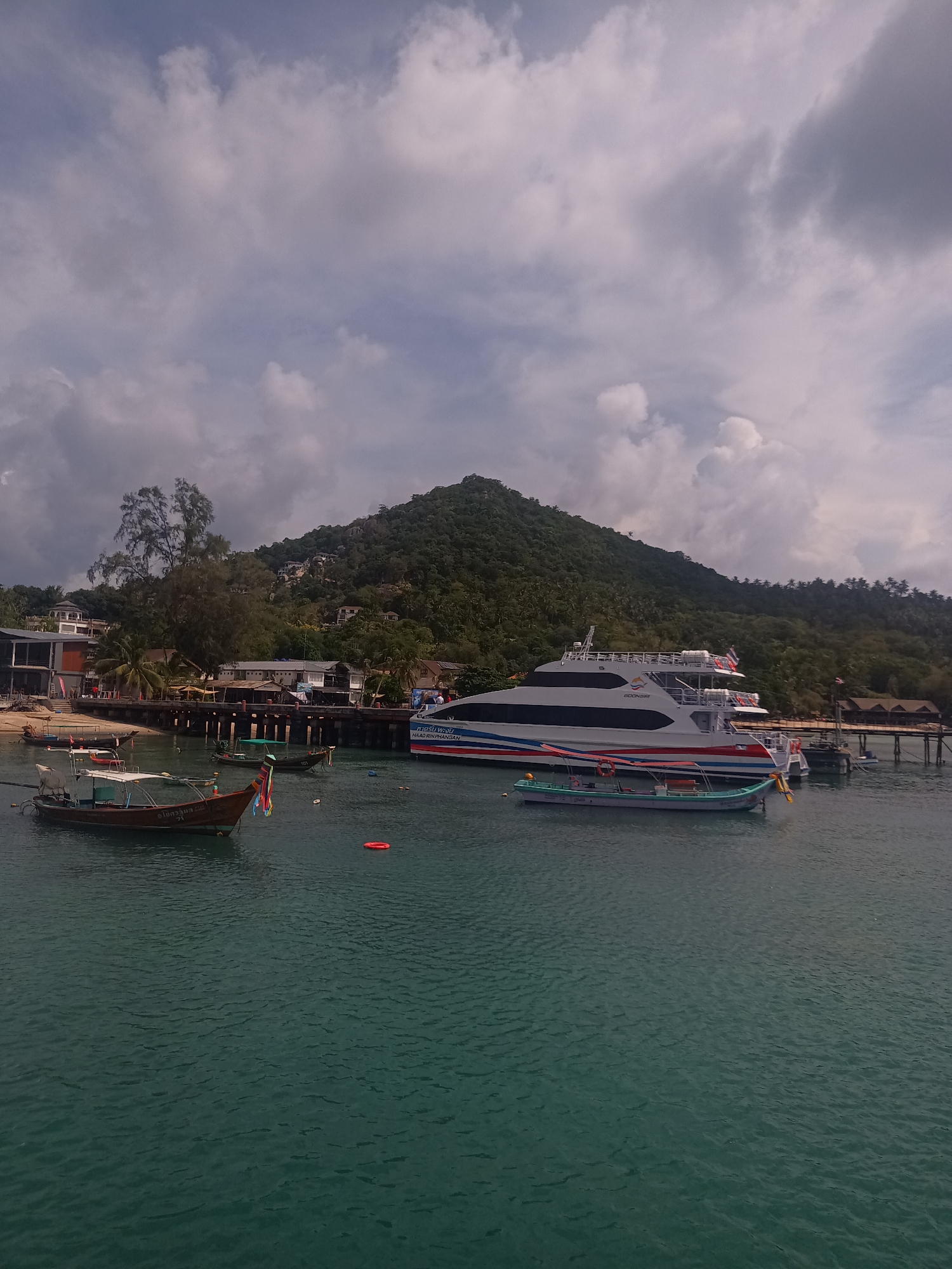 The image depicts a coastal scene in Tambon Ko Tao, Thailand. Dominating the view is a large, multi-story white ferry boat docked alongside a pier. Several smaller traditional Thai longtail boats are scattered in the foreground, some moored nearby. The water appears turquoise-green. A green, forested hill rises in the background, partially obscuring the sky which is filled with scattered, partly cloudy, grey-tinged clouds. Buildings are visible on the shore alongside the pier. An orange life preserver floats in the water.