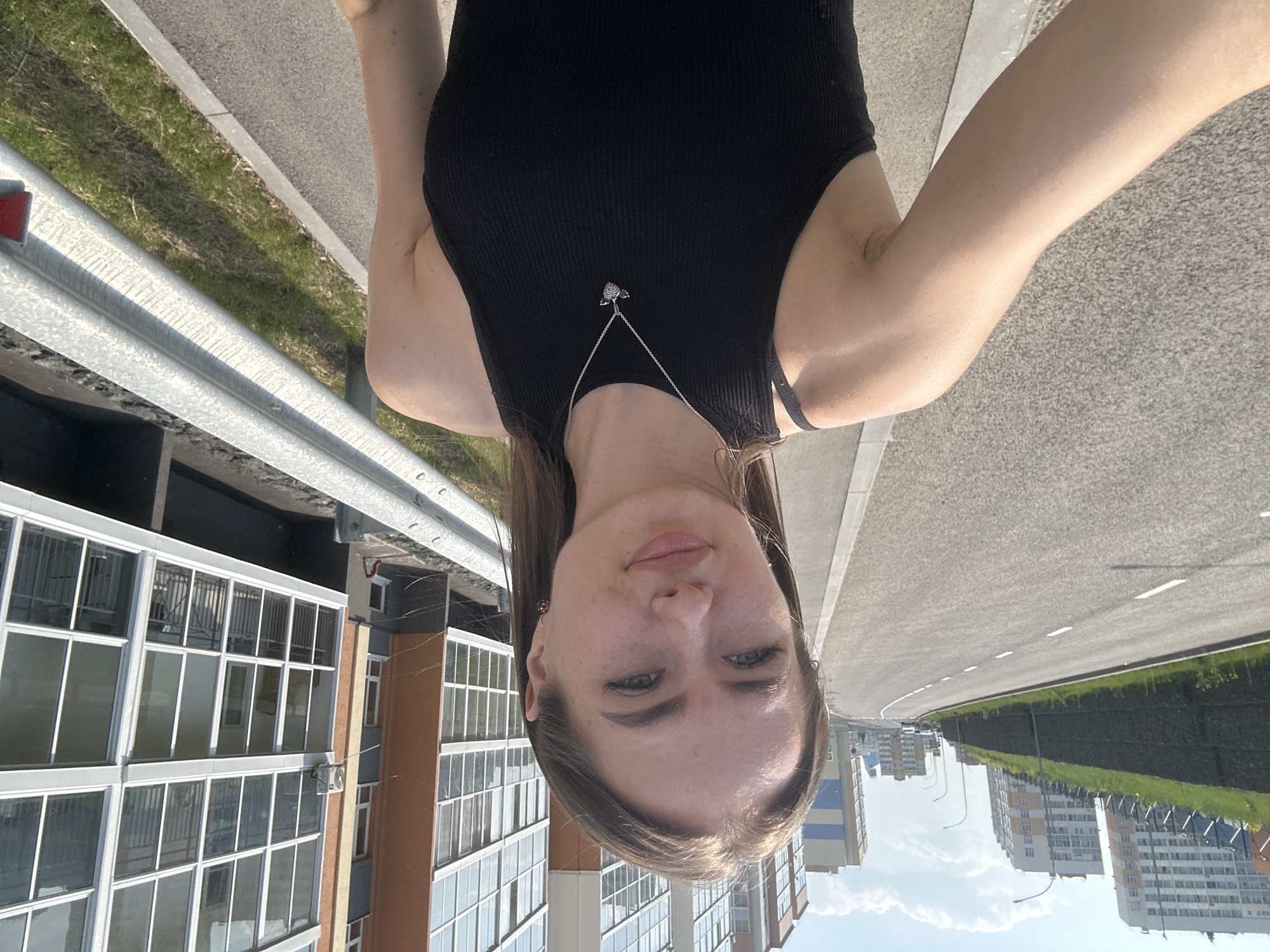 An outdoor, medium shot shows a young, fair-skinned woman with brown hair taking a selfie. She is wearing a black tank top with thin straps and a silver necklace with a heart-shaped pendant. Her hair is shoulder-length and she has light makeup on. She is standing on a paved sidewalk next to an asphalt road with white lines. In the background, there are tall, modern apartment buildings with many windows and balconies, a retaining wall with grass on top, and a metal guardrail. The sky is blue with some white clouds. The lighting suggests it is daytime.