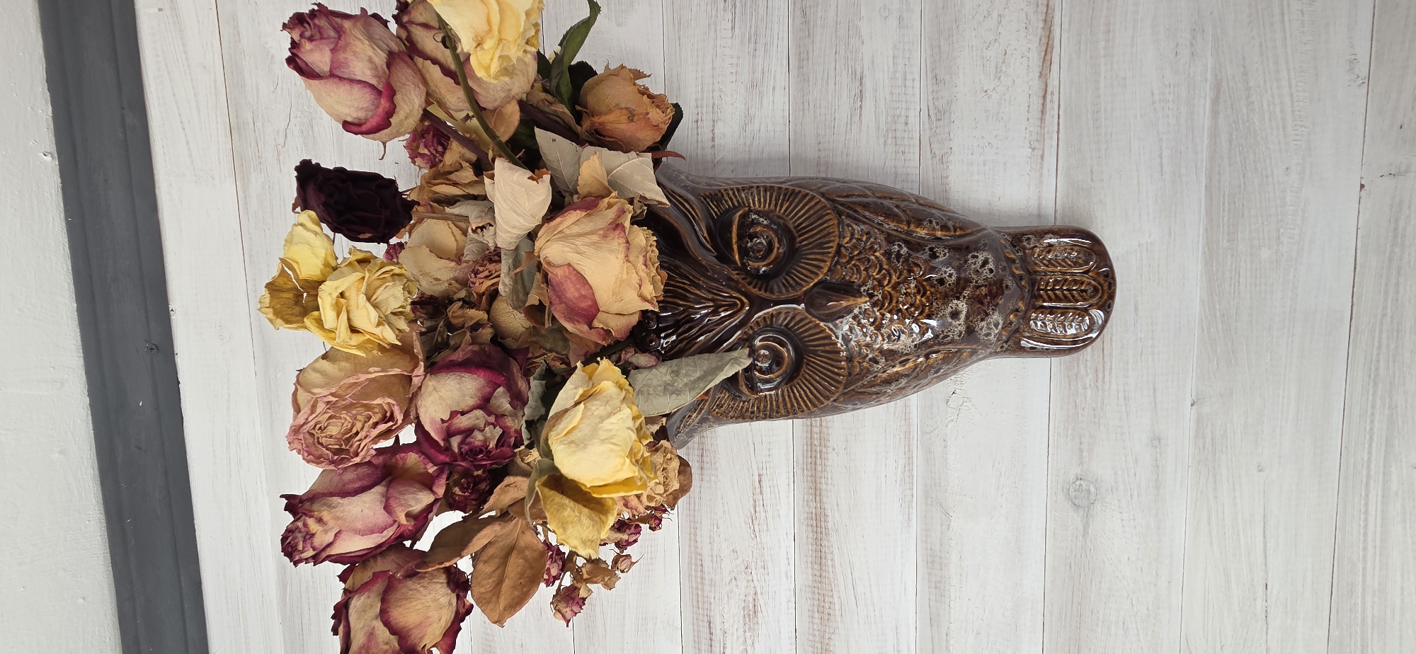 The image captures a still life arrangement featuring a decorative ceramic vase filled with dried flowers, set against a light-colored wooden background. The central object is a glossy, dark brown ceramic vase, intricately molded to resemble an owl. It displays textured details such as prominent, circular eyes with radial patterns and scaled or feathered body sections, with varying shades of brown and speckled glazes.
Emerging from the vase is a bouquet of dried roses. These flowers exhibit a range of faded colors, primarily deep maroon, pale yellow, and various shades of beige and light brown, indicative of their dried state. The petals appear shriveled and brittle, and some dried leaves are also visible within the arrangement. The overall impression of the flowers is one of natural decay, lending a vintage or rustic aesthetic to the composition.
The background consists of light-toned wooden planks, likely a table or floor, oriented vertically in the frame. On the far left, two distinct darker vertical lines, possibly shadows or part of a structural element, break up the otherwise uniform wooden surface. There is no visible activity or event taking place, and no people are present. The lighting is soft and even, highlighting the textures of the ceramic and the delicate, aged petals. No text is visible in the media file.