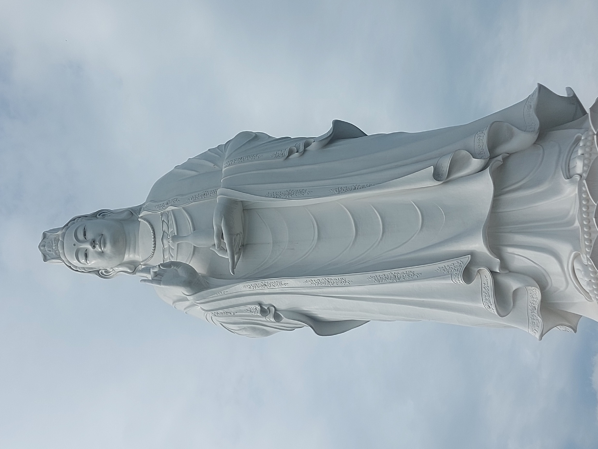 This is a low-angle, eye-level, full-shot photograph of a large, white, stone statue of Guanyin (also known as Quan Âm in Vietnam) standing against a pale, overcast sky. The statue depicts her in traditional robes, with her right hand raised in a gesture of blessing and her left hand holding a small vase. The statue is intricately detailed, with flowing drapery and delicate ornamentation. The statue is located at the Linh Ứng Pagoda on Sơn Trà Peninsula in Đà Nẵng, Vietnam, a prominent landmark and tourist attraction. The overall mood of the image is serene and majestic.