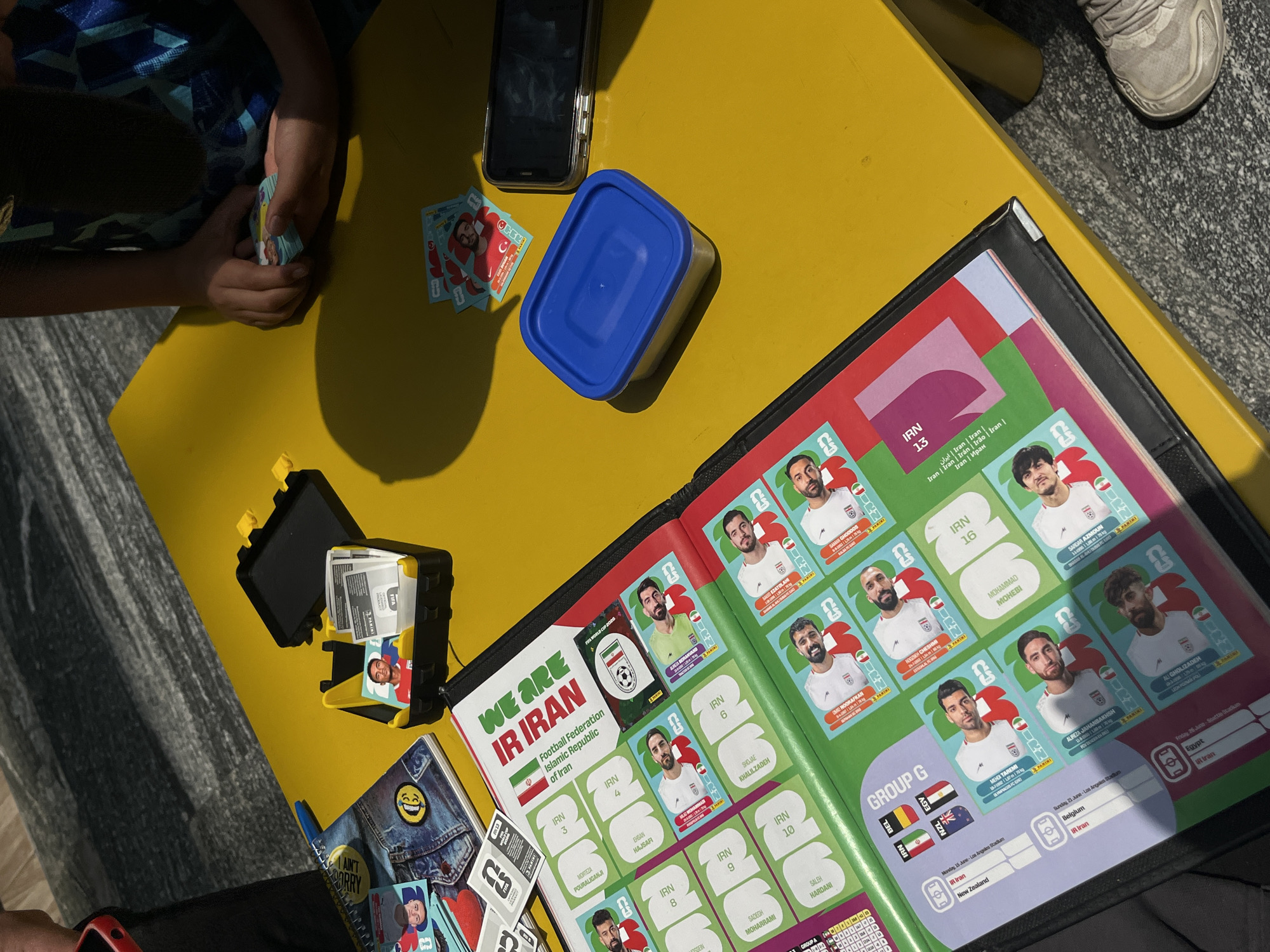 The image shows a person sitting at a yellow table, sorting and organizing Panini World Cup stickers. The stickers are of the Iranian national team. A sticker album, a blue container with a lid, a phone, and a sticker organizer are on the table. The person appears to be placing stickers into the album. A shoe is visible at the upper right corner of the image.