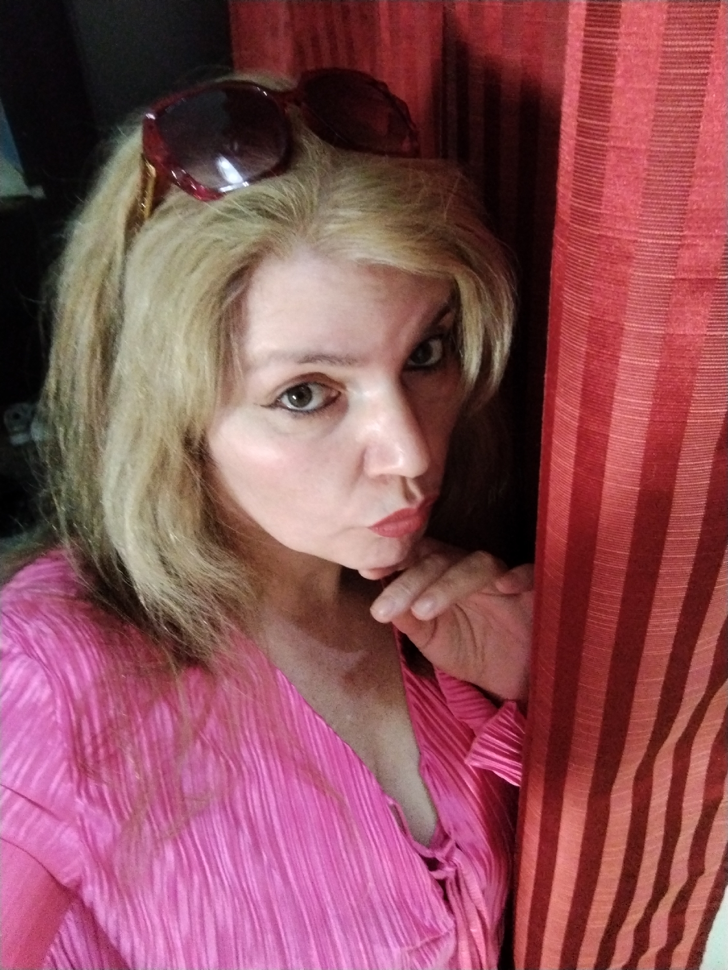 A woman with blonde hair and sunglasses perched on her head poses indoors. She is wearing a pink shirt with a V-neck. Her left hand is near her face, and she is looking directly at the camera with a slight pout. Part of a red, striped curtain is visible on the right side of the frame. The location is uncertain, but the request mentioned Al Mas'oudi, United Arab Emirates, which may or may not be relevant.