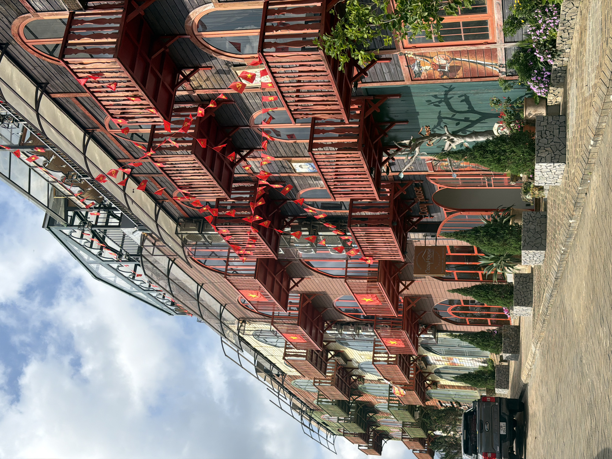 The image shows the exterior of a building in Măng Đen, Vietnam. The building has multiple levels with a distinctive architectural style, featuring red balconies decorated with Vietnamese flags. The facade incorporates wood and other materials, creating a unique aesthetic.
A sidewalk is visible in the foreground, along with parked cars. The sky above is partly cloudy. Lush greenery surrounds the building.