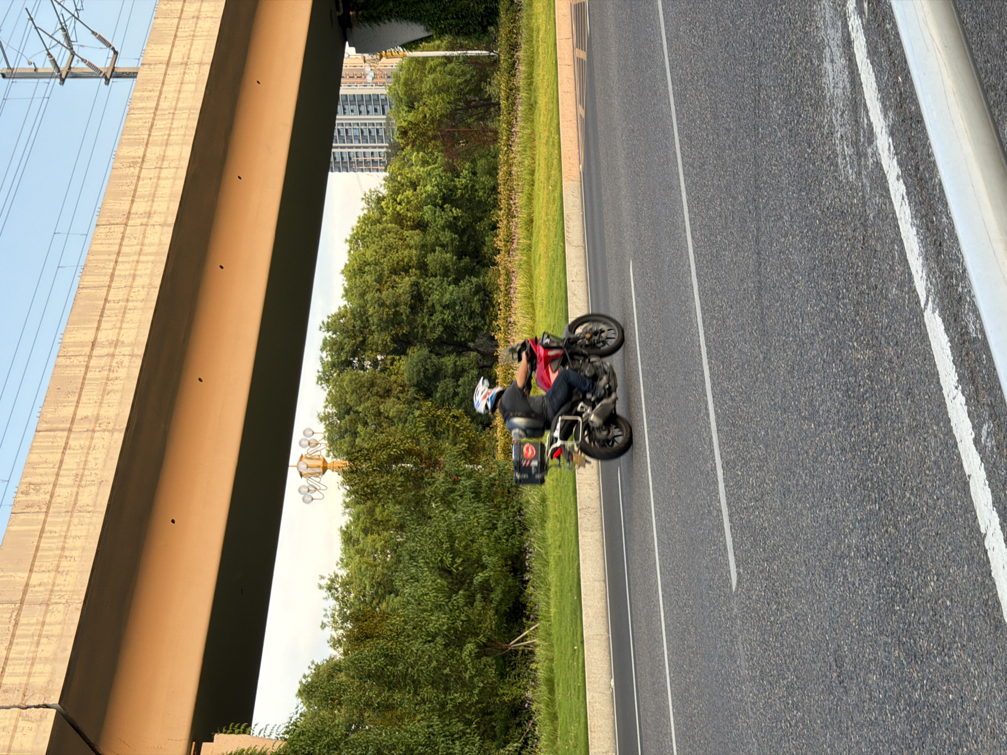 A person wearing a helmet is riding a red motorcycle with a black cargo box on the back. The motorcycle is on an asphalt road with white lane markings. The road is bordered by a grassy median with some small purple flowers. Behind the median, there is a dense line of trees. In the background, there are tall buildings and a bridge structure. The scene appears to be in the city of Jiangjin, China, based on the context. The sun is casting long shadows, suggesting it is either early morning or late afternoon.